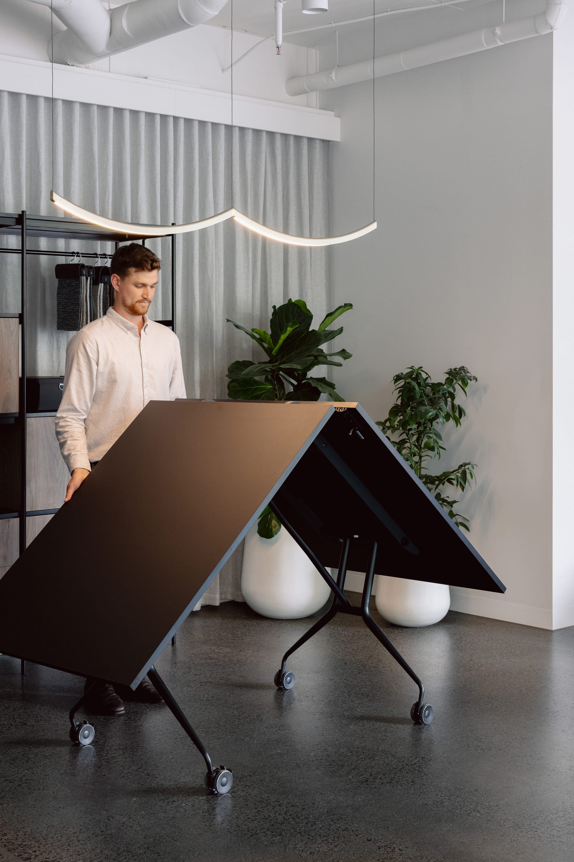 Libro Folding Table by Mara SRL. Black table in a pitched position as it is being folded by man with a white shirt, in space with dark concrete floors and white potted plants. 