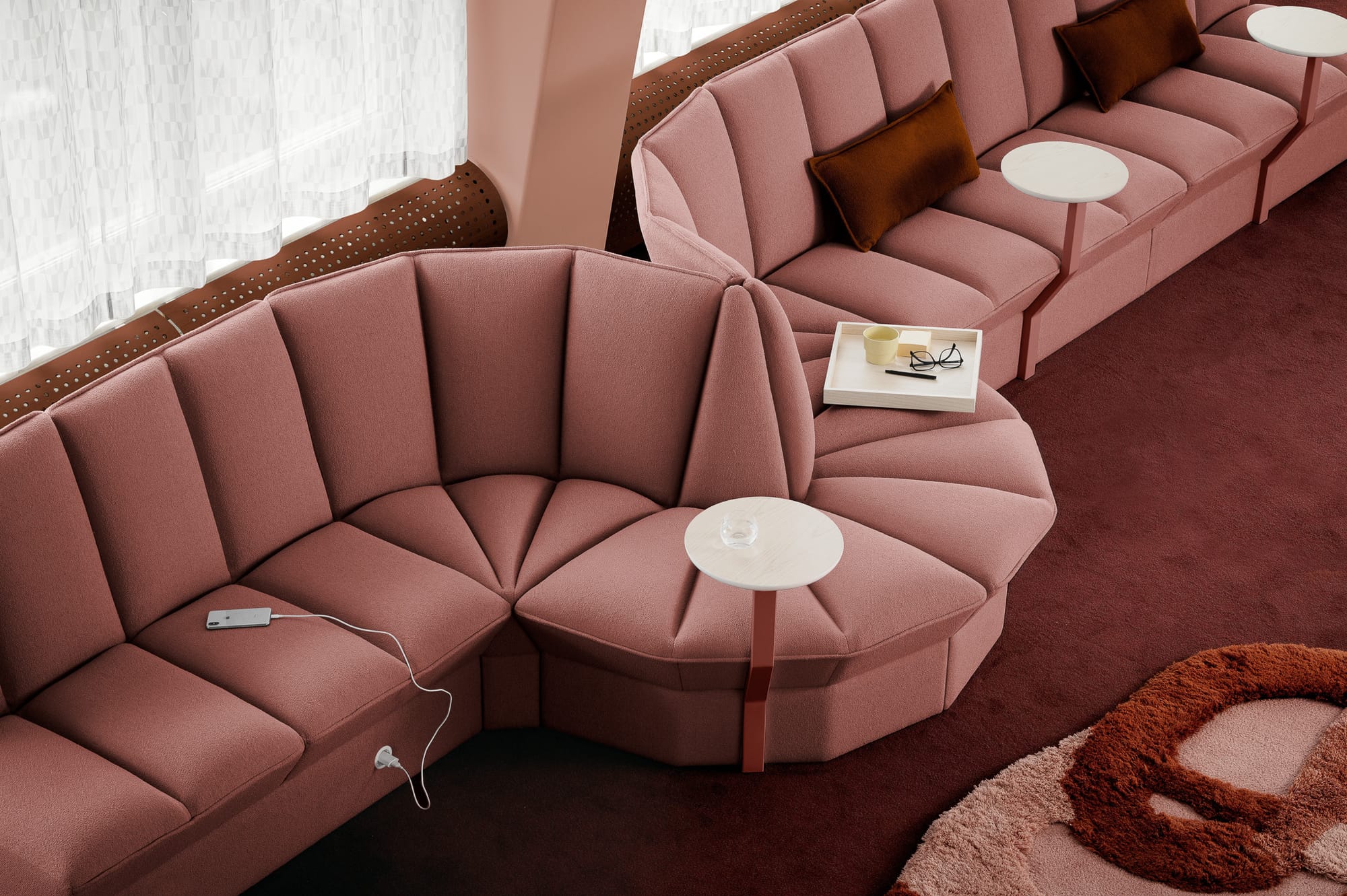 Ori Lounge by EFG. Curved and tufted salmon couch with integrated side tables at various points in space with white curtains and red carpet. 