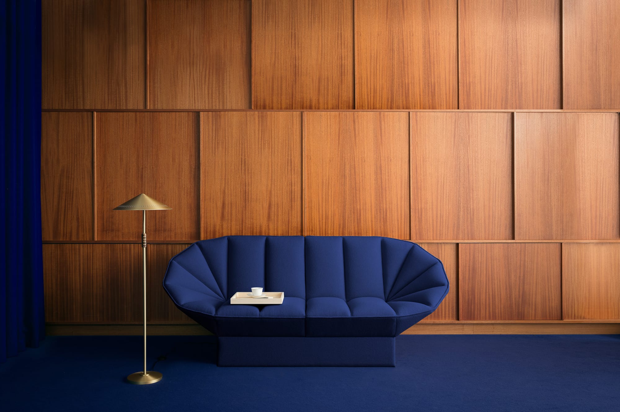 Ori Lounge by EFG. An electric blue couch with curved edges and tufting in space with timber clad wall panels, electric blue carpet and a gold floor lamp. 