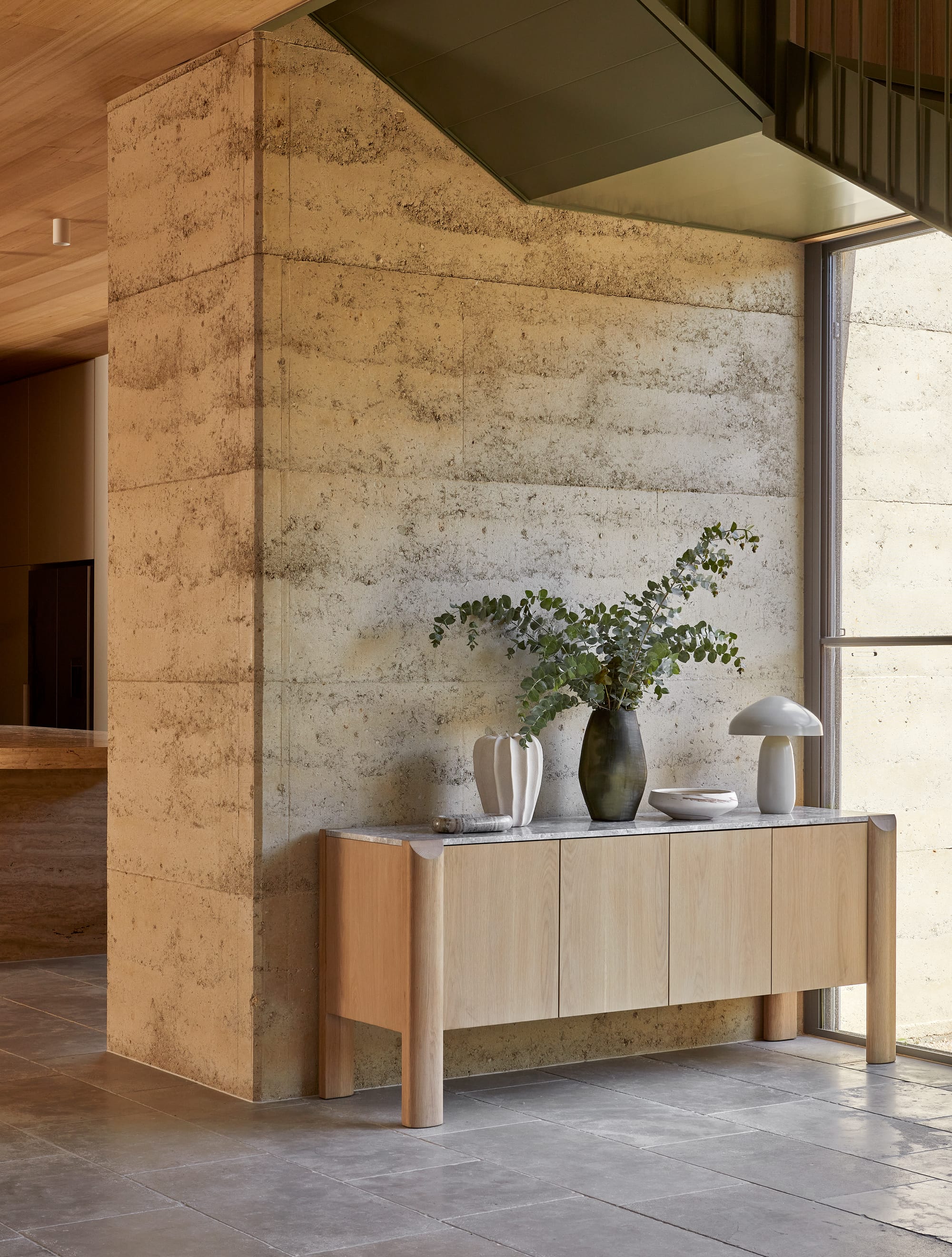 Willa by GlobeWest from Collection 2025.  Light wood sideboard with dark planted pot on top, in front of rammed earth wall.