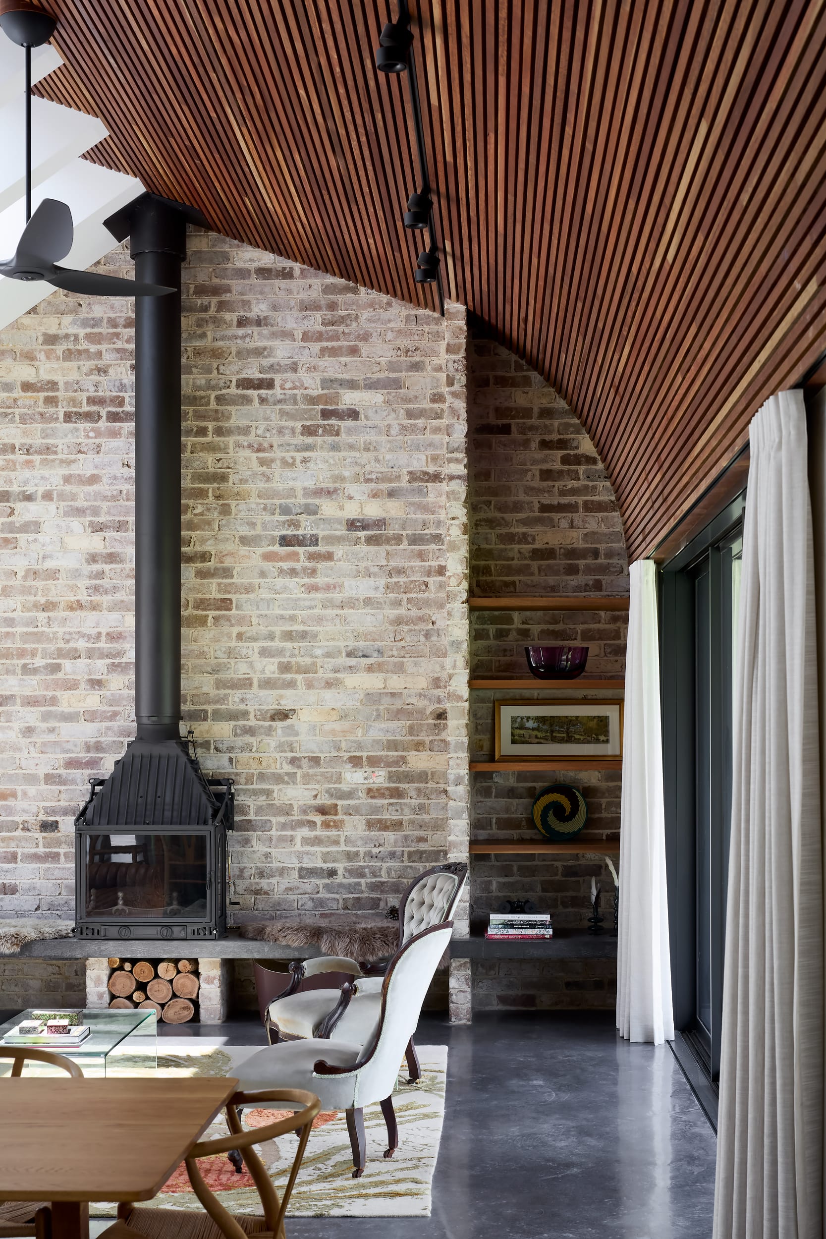 House Kimberley by AO Design Studio. Photography by Luc Remond. Living space with recycled brick walls, concrete floors, curved timber clad ceiling and black fireplace. Eclectic and vintage inspired furnishings in space. 