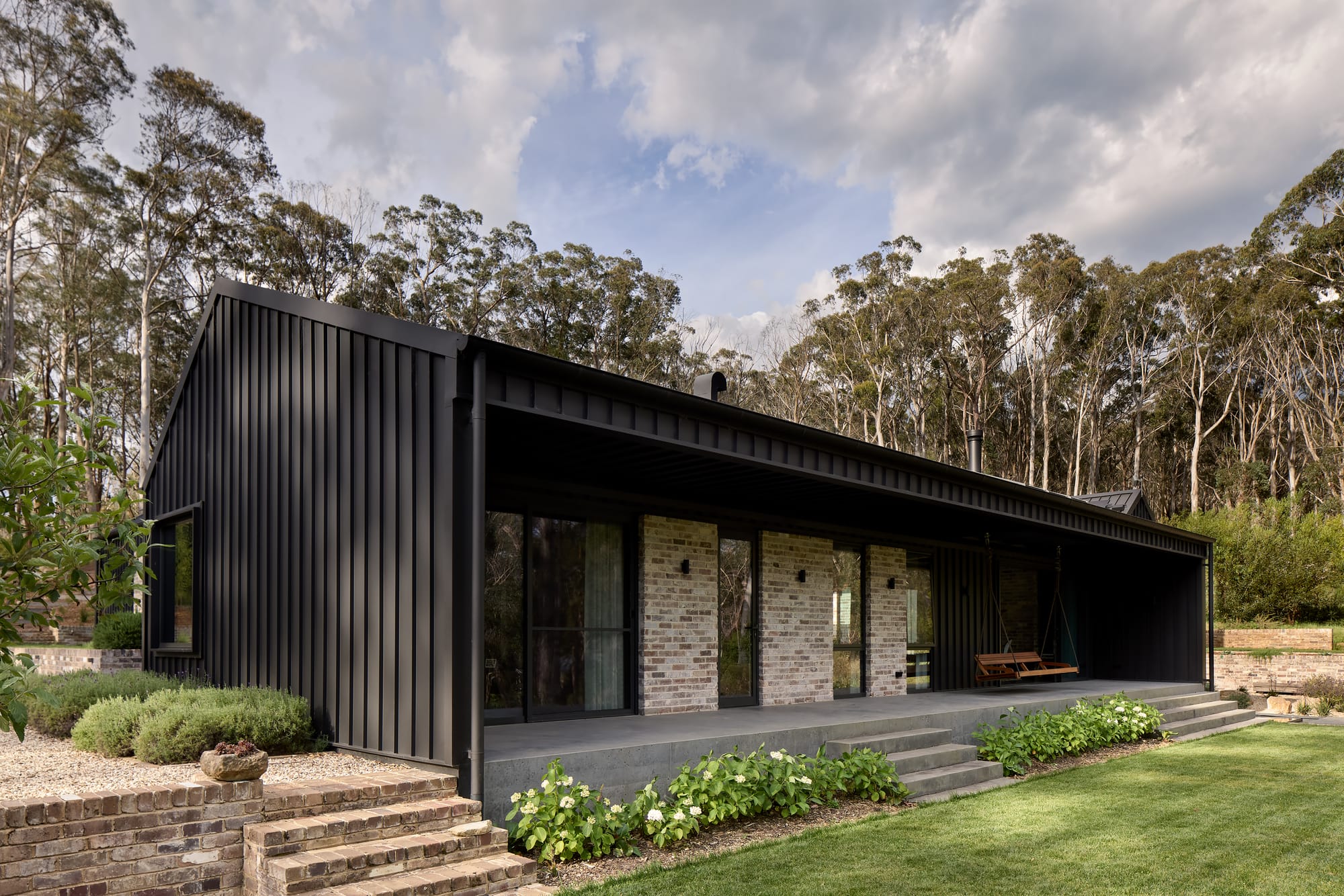 House Kimberley by AO Design Studio. Photography by Luc Remond. Pavilion style home with black clad exterior, concrete patio and steps, recycled brick feature walls and long stretch of green grass. 