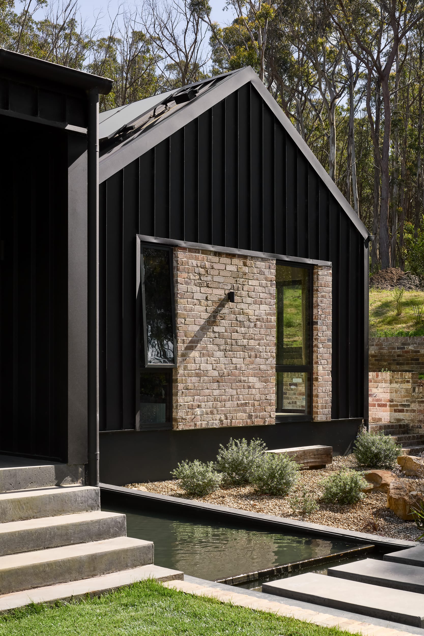 House Kimberley by AO Design Studio. Photography by Luc Remond. Pavillion style black clad exterior of home with recycled brick details, concrete stairs and pathways across integrated water features. 