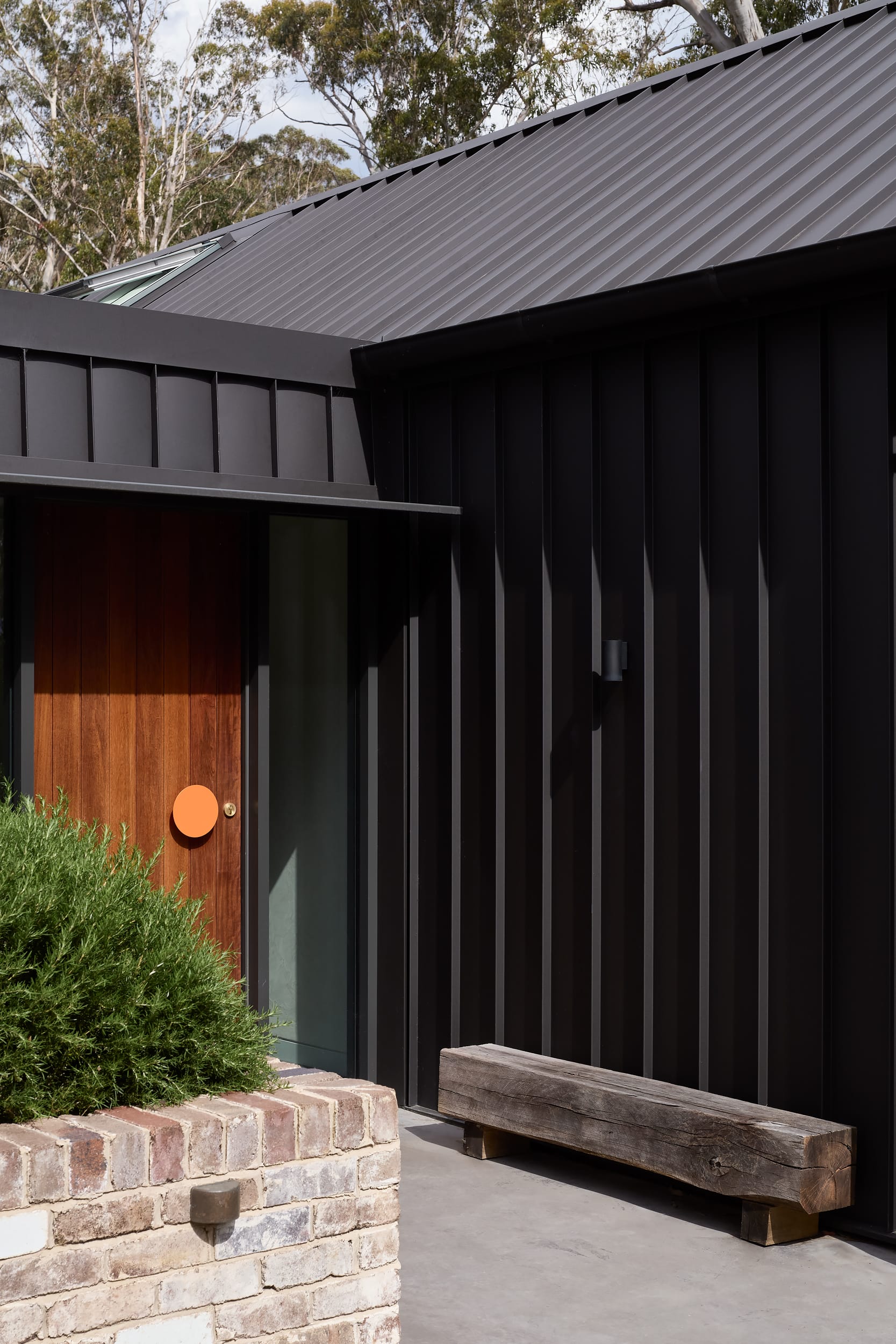 House Kimberley by AO Design Studio. Photography by Luc Remond. Timber front door with pale orange round door handle. Black clad exterior walls with recycled brick garden bed, and recycled timber bench seat by door. 