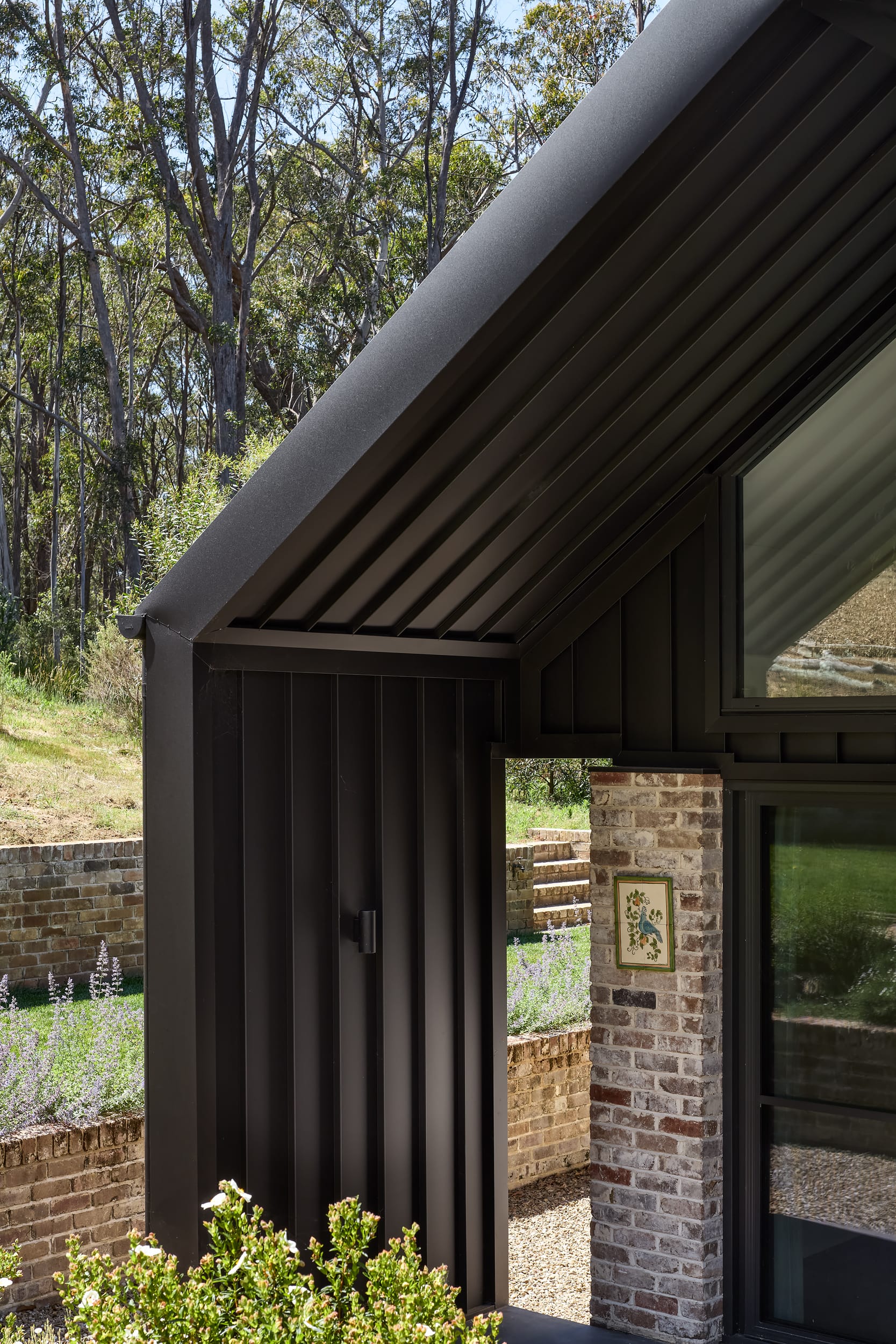 House Kimberley by AO Design Studio. Photography by Luc Remond. Black clad external roof arch next to recycled brick pillar and garden bed. 