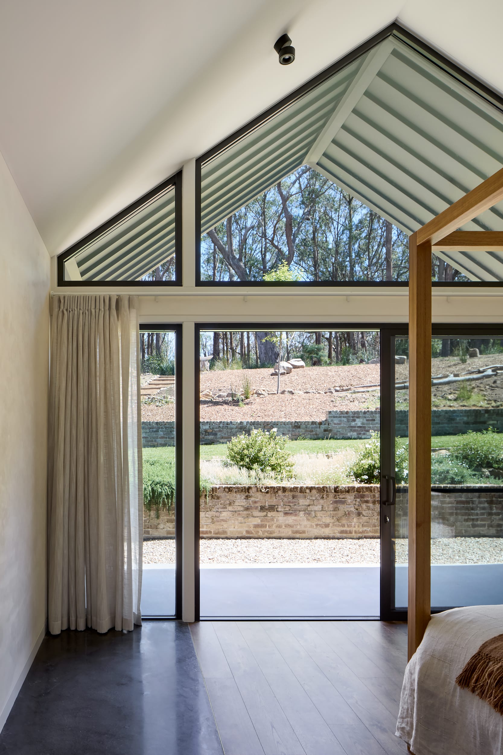 House Kimberley by AO Design Studio. Photography by Luc Remond. Pitched window and ceiling in bedroom, opening onto landscapes garden with concrete patio, recycled brick garden beds, green lawn and gravel. 