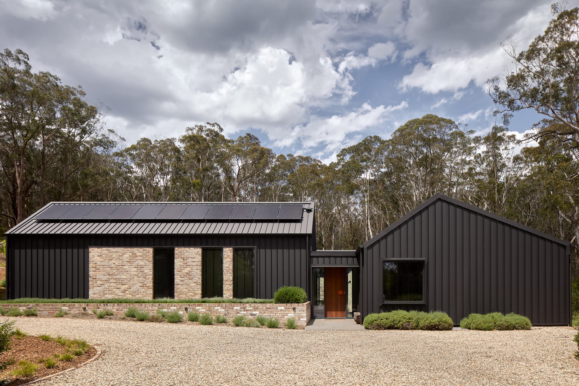 House Kimberley by AO Design Studio. Photography by Luc Remond. Front facade of black clad pavilion home with pale gravel driveway, recycled brick feature walls and garden beds. 