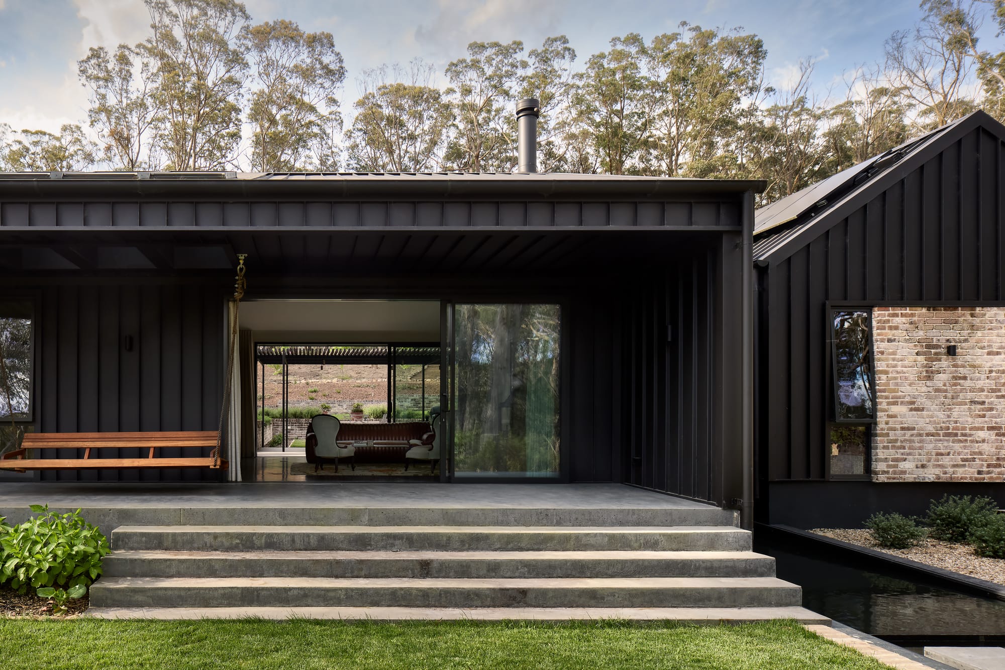 House Kimberley by AO Design Studio. Photography by Luc Remond. Concrete vernadah and steps leading from green grass to timber clad home with large glass sliding doors. 