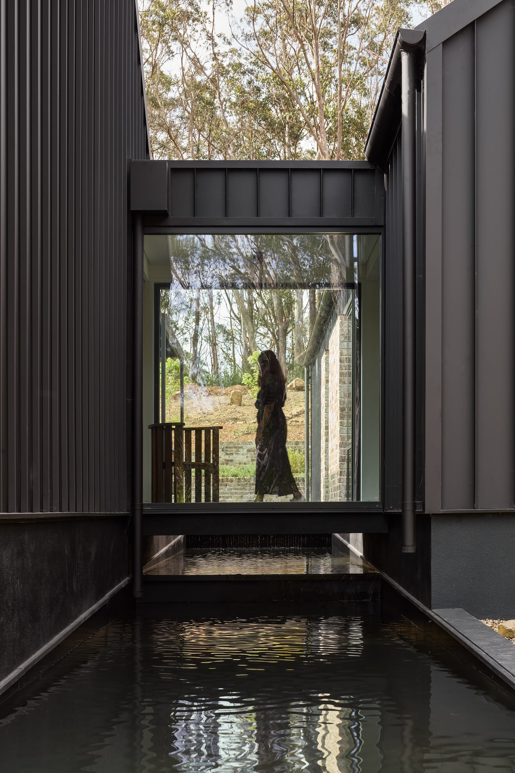 House Kimberley by AO Design Studio. Photography by Luc Remond. Water running underneath elevated glass walkway finished with black exterior clad and framing gardens with native Australian plants. 