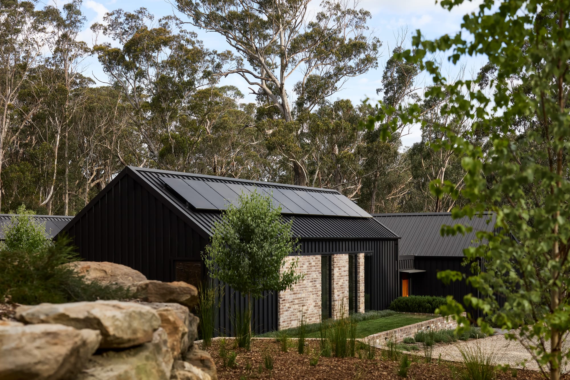 House Kimberley by AO Design Studio. Photography by Luc Remond. Black clad home with pitched rood and recycled brick feature walls, with tired gardens outlined in recycled brick and featuring green grass and fruit trees. 