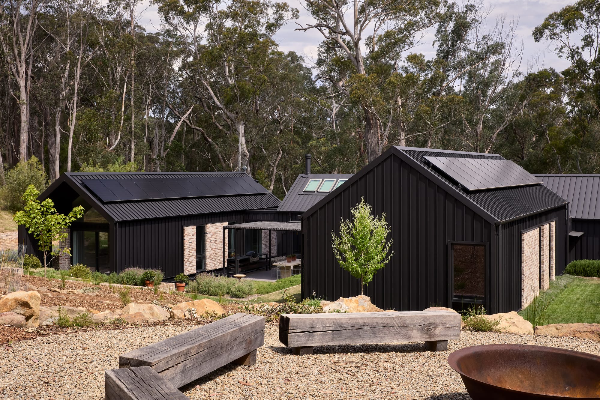 House Kimberley by AO Design Studio. Photography by Luc Remond. Black clad pavillion style home with distinct wings arranged in background of image and gravel area in foreground, with recycle timber bench seating arranged around a rusted firepit. 