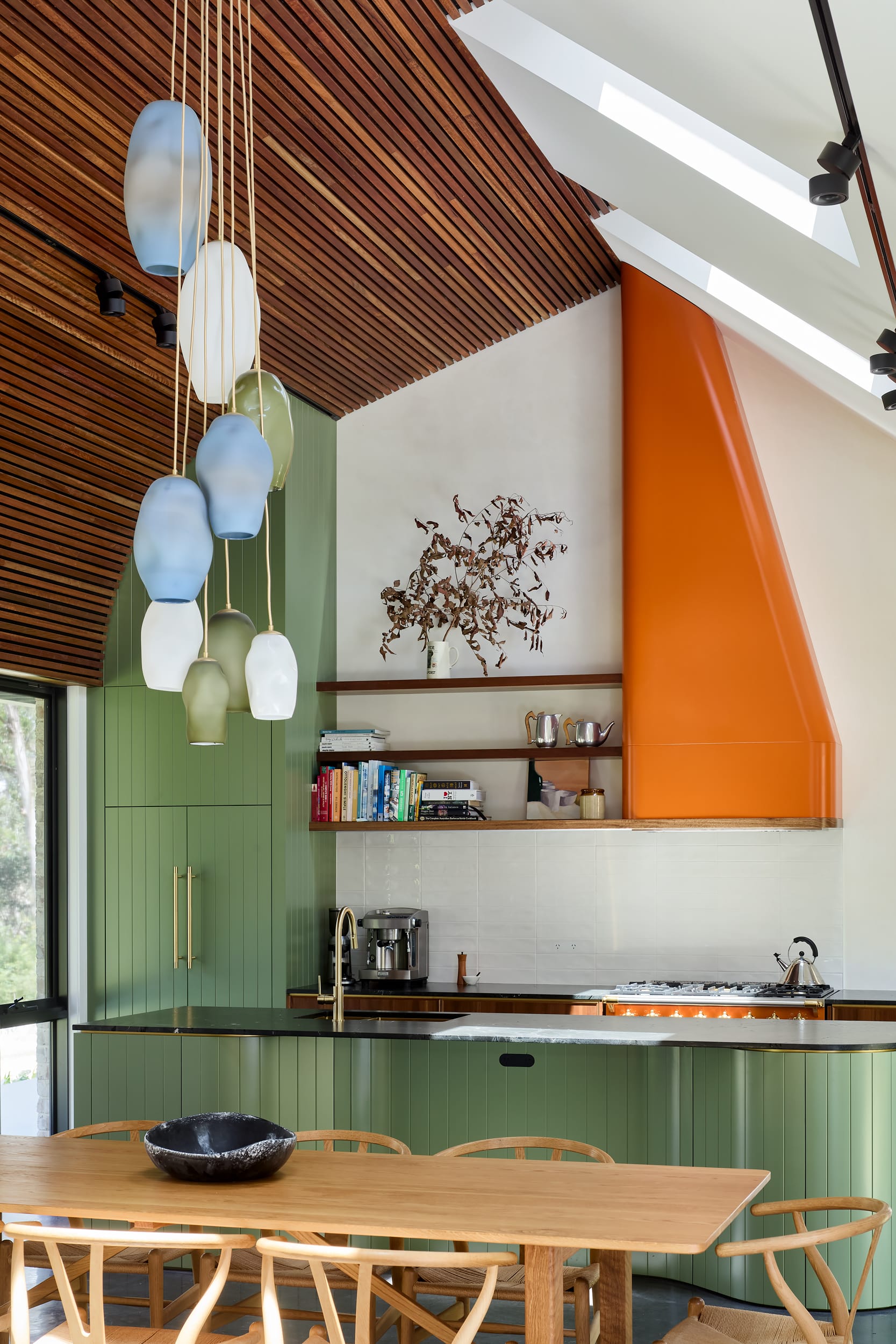 House Kimberley by AO Design Studio. Photography by Luc Remond. Green timber kitchen with orange accent venting, floating timber benches and a timber dining table in the foreground. 