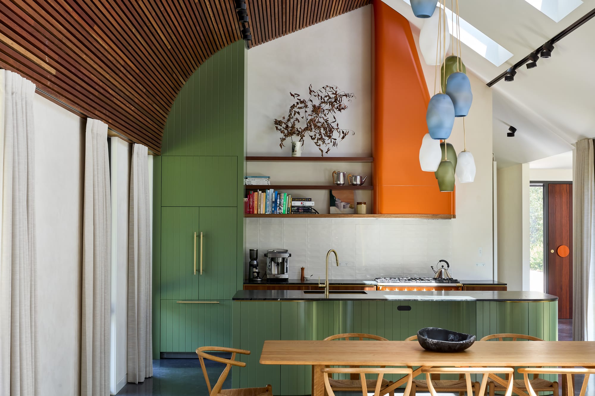 House Kimberley by AO Design Studio. Photography by Luc Remond. Green timber kitchen with orange accent induction vent and timber floating shelves. Timber dining table with green, blue and white abstract pendant light hanging above. 