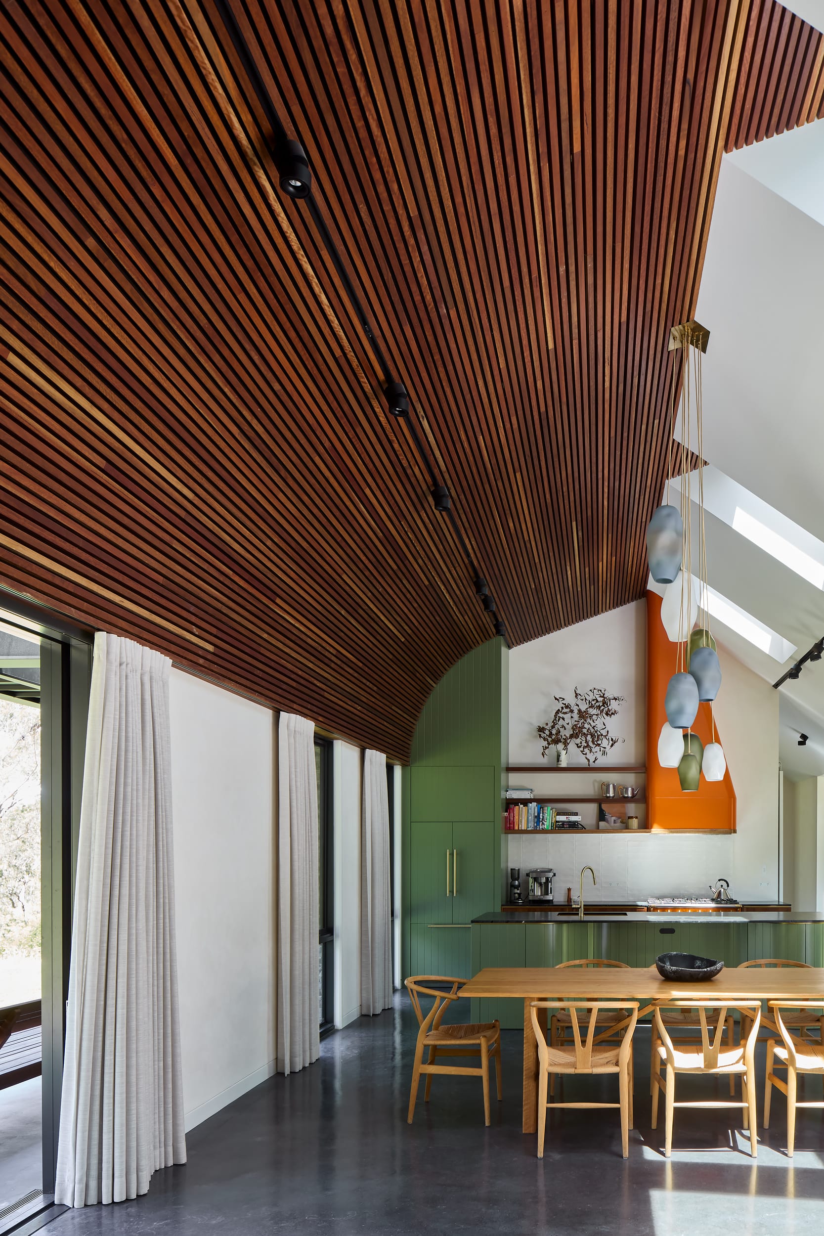 House Kimberley by AO Design Studio. Photography by Luc Remond. Long open plan living space with curved timber clad feature ceiling, green and orange kitchen, timber dining table and black polished concrete floors. 