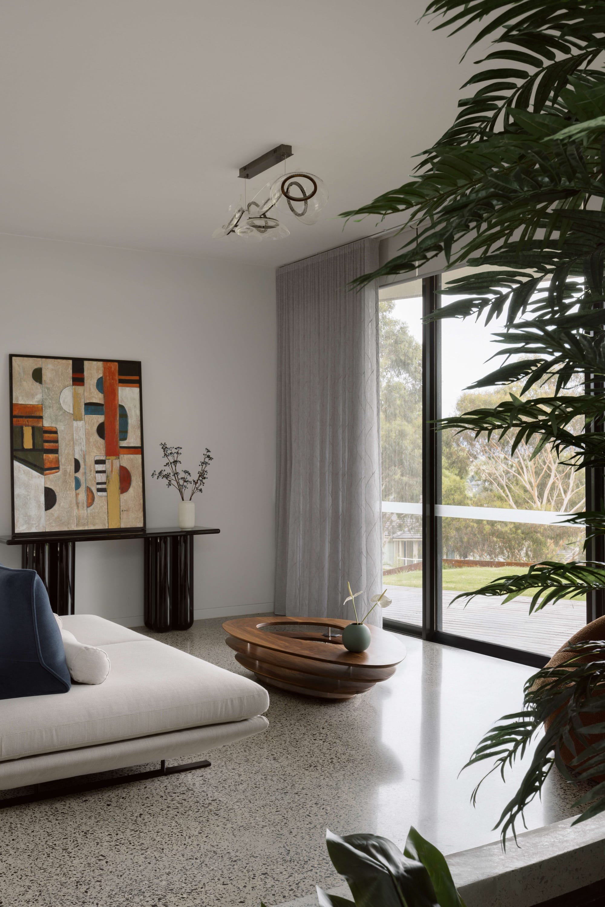 House On The Hill by ENCLAVE ARCHITECTS. Photography by Elise Scott. Living space with polished concrete floors, white couch, round tiered timber coffee table, leading to an outdoor timber deck overlooking grassy landscape.