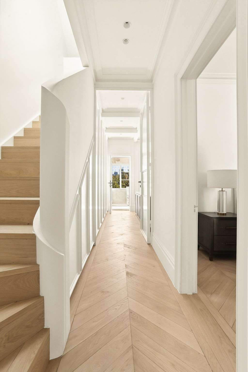 Glenmore Road House by BY SOMA. Photograph by Daniel Cook. Hallway with blonde herringbone timber flooring, white walls, a curved staircase and a view down the hall to a kitchen and onto a garden.