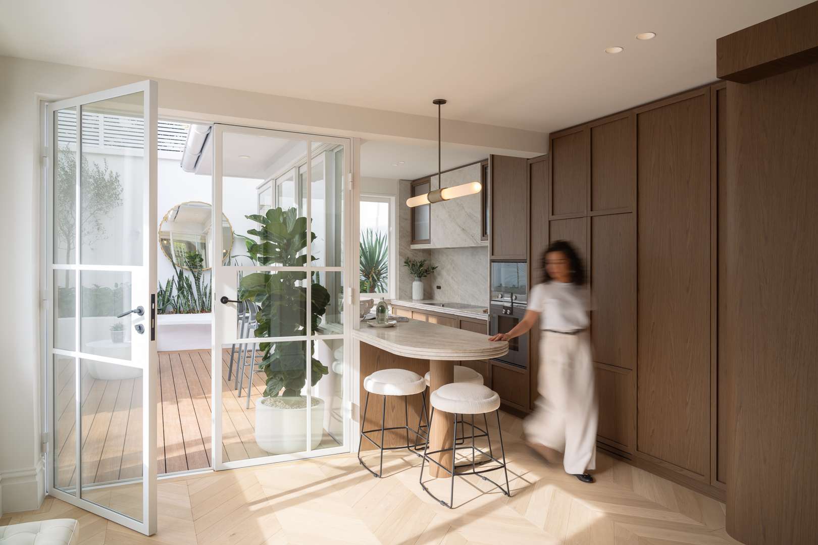 Glenmore Road House by BY SOMA. Photograph by Daniel Cook. Kitchen with herringbone blonde timber floors, warm curved timber cabinetry, white grid doors and windows opening onto a deck.