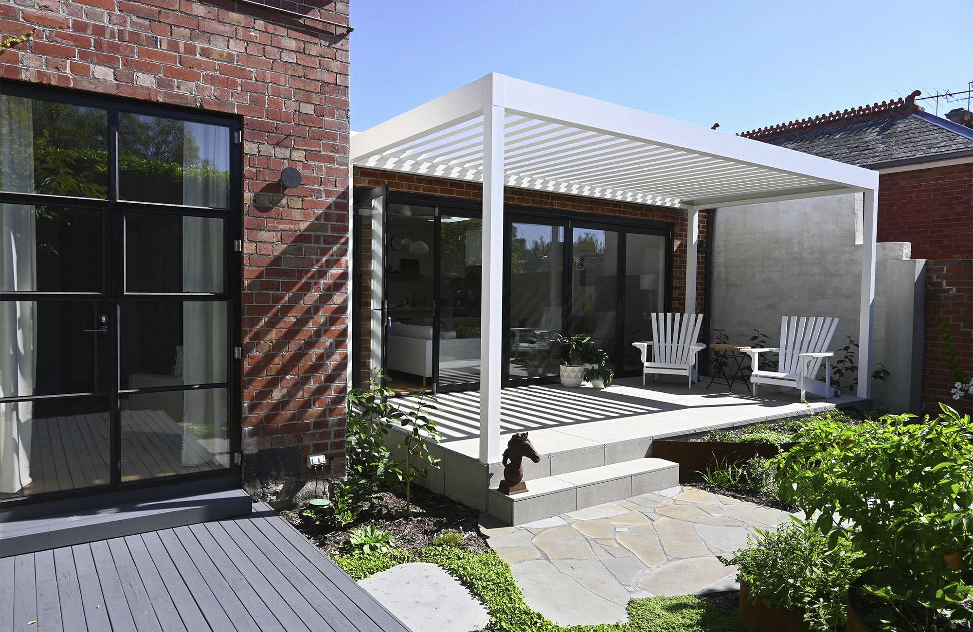 Scandi House by SG2 architects + interiors. Photography by Sebastiano Ghezzi. Rear facade of single story residential home with two structures, one made from recycled brick with black grid windows and doors, and another white pergola structure leading down steps onto a paved courtyard garden.