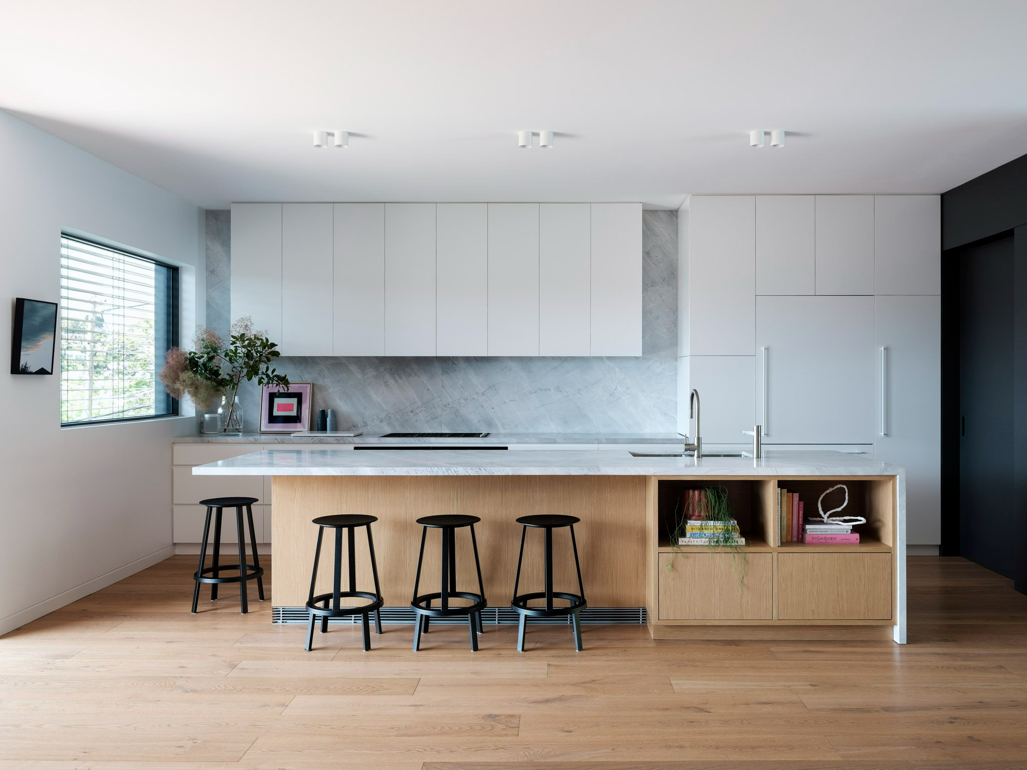 Minimbah by Castlepeake Architects. Photography by Tom Ross. Kitchen with light timber island with integrated storage, black bar stools, white cabinetry, light blue marbled splashback, white walls and timber floors.