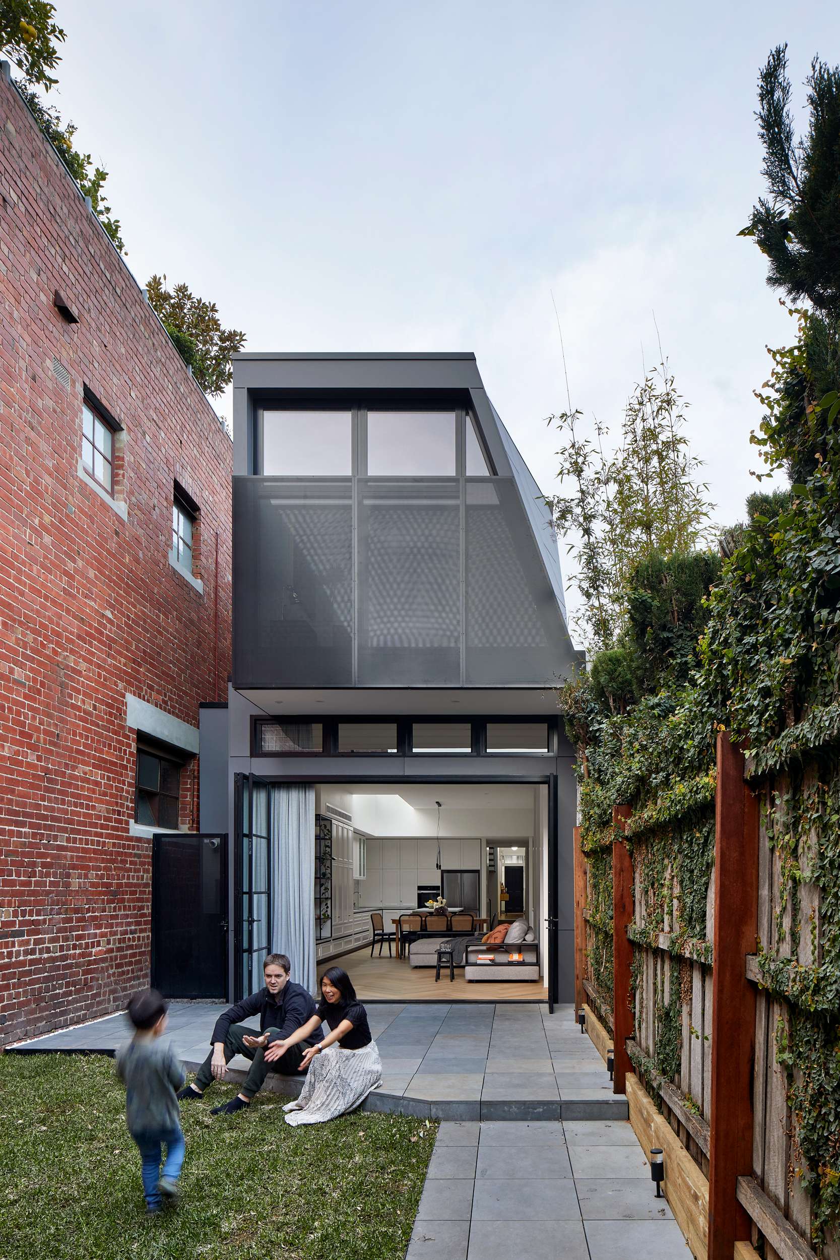 Richardson Middle Park by Chan Architecture. Photography by Tatjana Plitt. Rear facade and garden of multistorey home with full height folding doors opening back patio onto open plan living, dining and kitchen space.