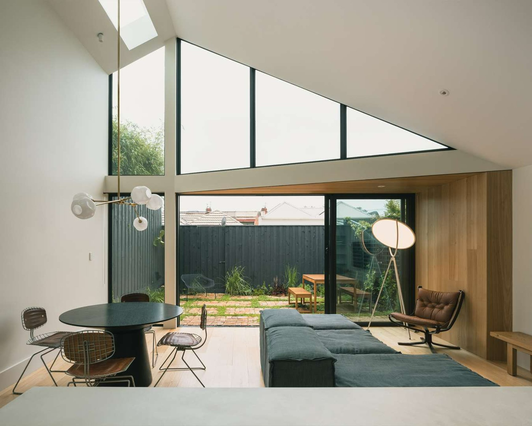 Abbotsford House by Eckersley Architects. Photography by Dan Preston. Ope plan living space with sloping ceilig and full height angular windows, with timber paneled feature walls, brown leather furnishings and a small brick courtyard.