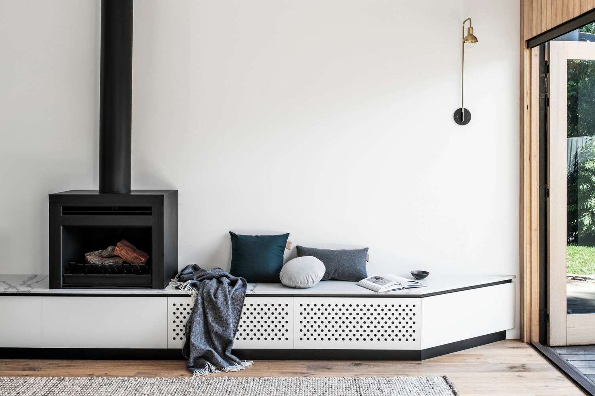 Silhouette Hytte House by FIGR Architecture. Photography by Tom Blachford. White wall with white integrated sideboard with perforated cupboard faces, with a black fireplace on top and pillows and throws in blue and navy tones.