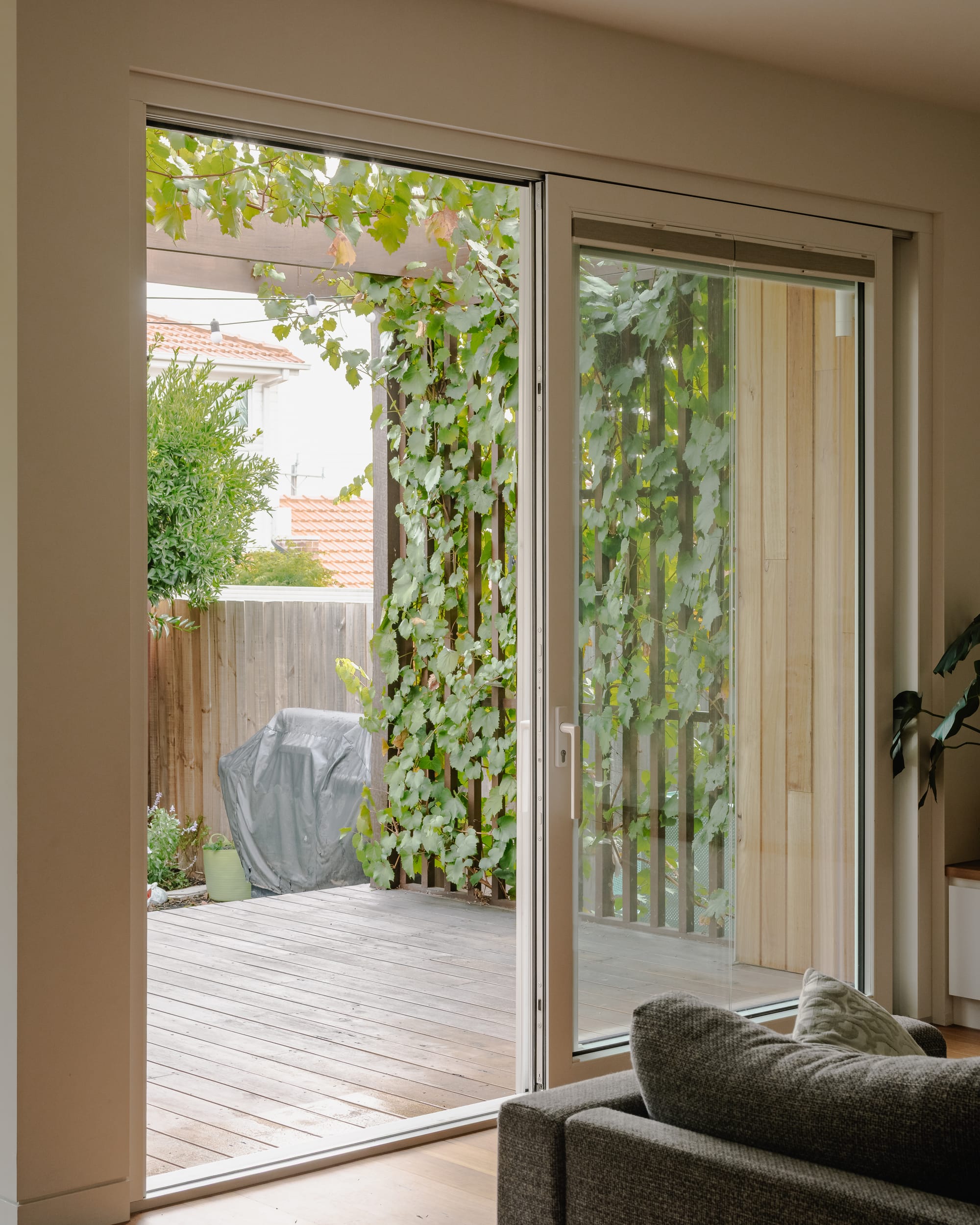 House in Preston by Ashley O'Neill Architects. Photography by Victor Vieaux. White framed glass sliding doors opening onto timber porch with vertical and horizontal timber pergola enclosing the space, covered in vine leaves.