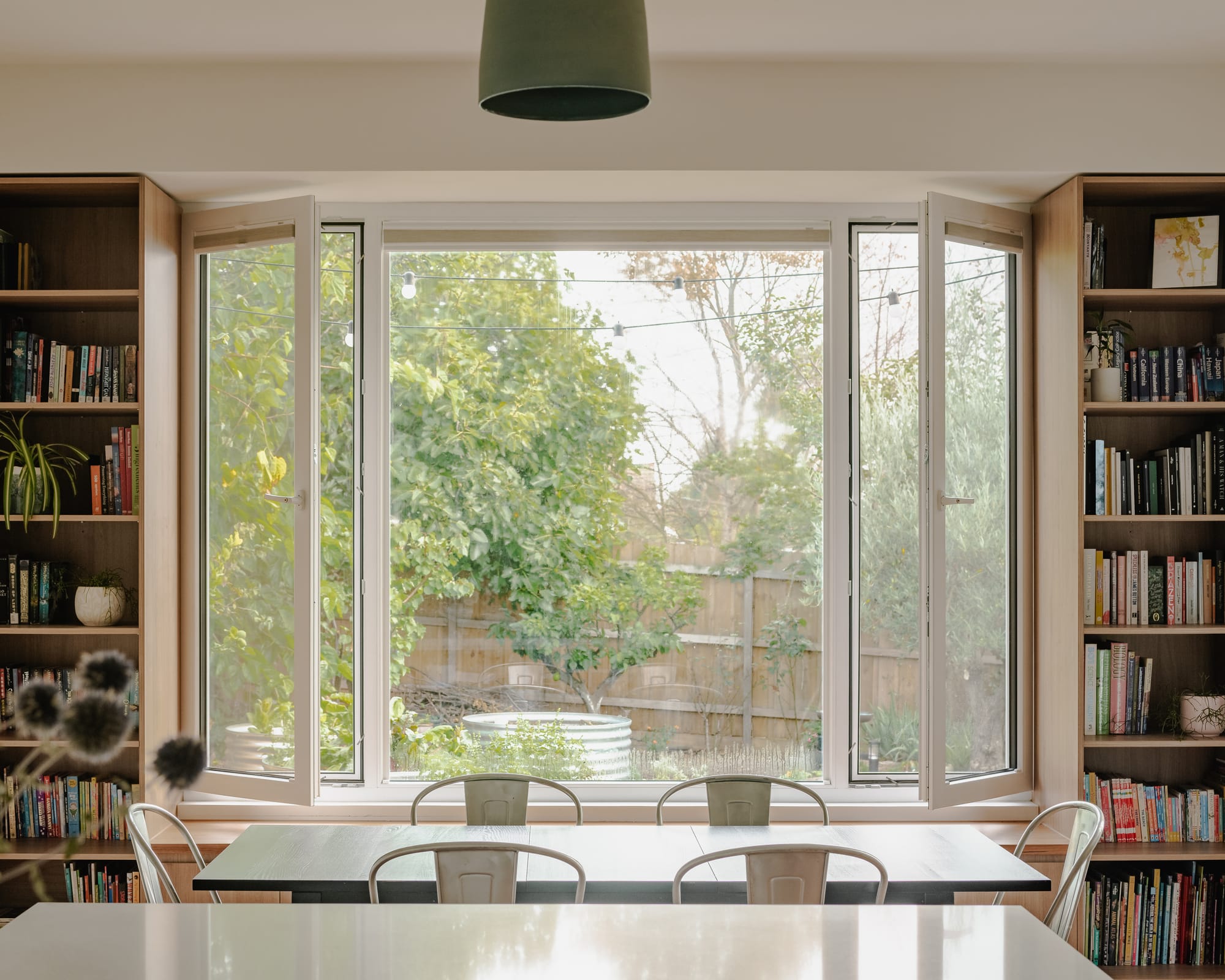 House in Preston by Ashley O'Neill Architects. Photography by Victor Vieaux. Dining table with chrome chairs, positioned in front of a low timber bench and timber bookshelves to the left and right of a white framed window, opening up onto the garden below.
