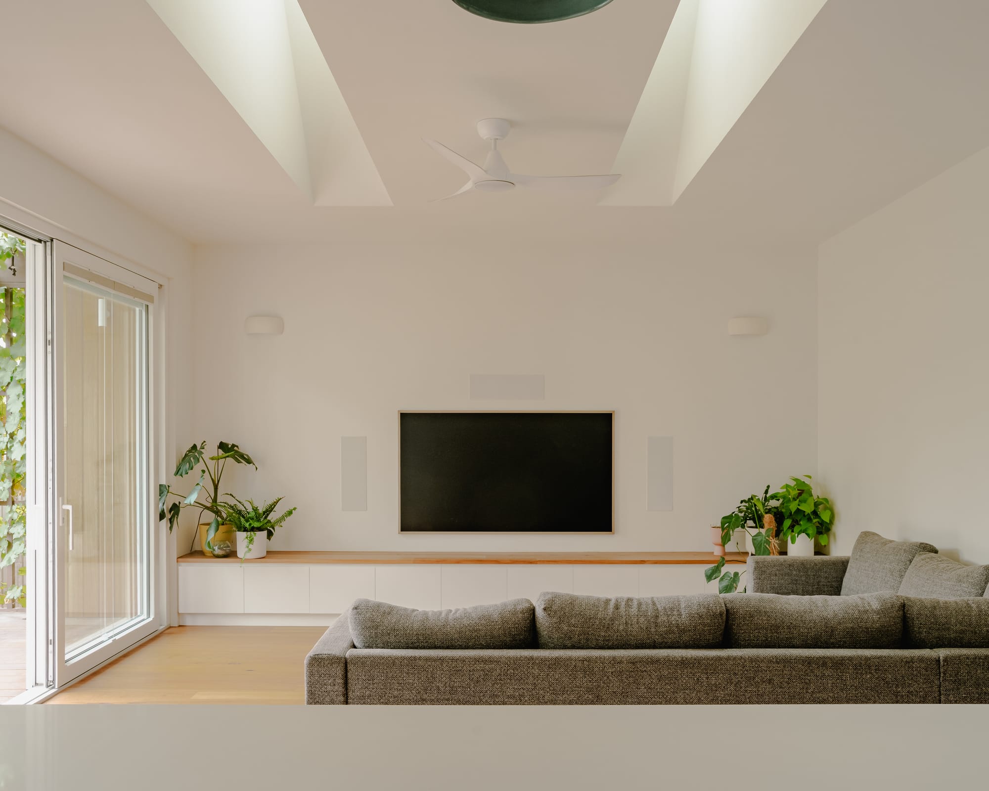 House in Preston by Ashley O'Neill Architects. Photography by Victor Vieaux. Living space with a grey, modular lounge, a hall-hung flat screen TV, integrated sideboard with white cabinet faces and a timber top running the length of the space.