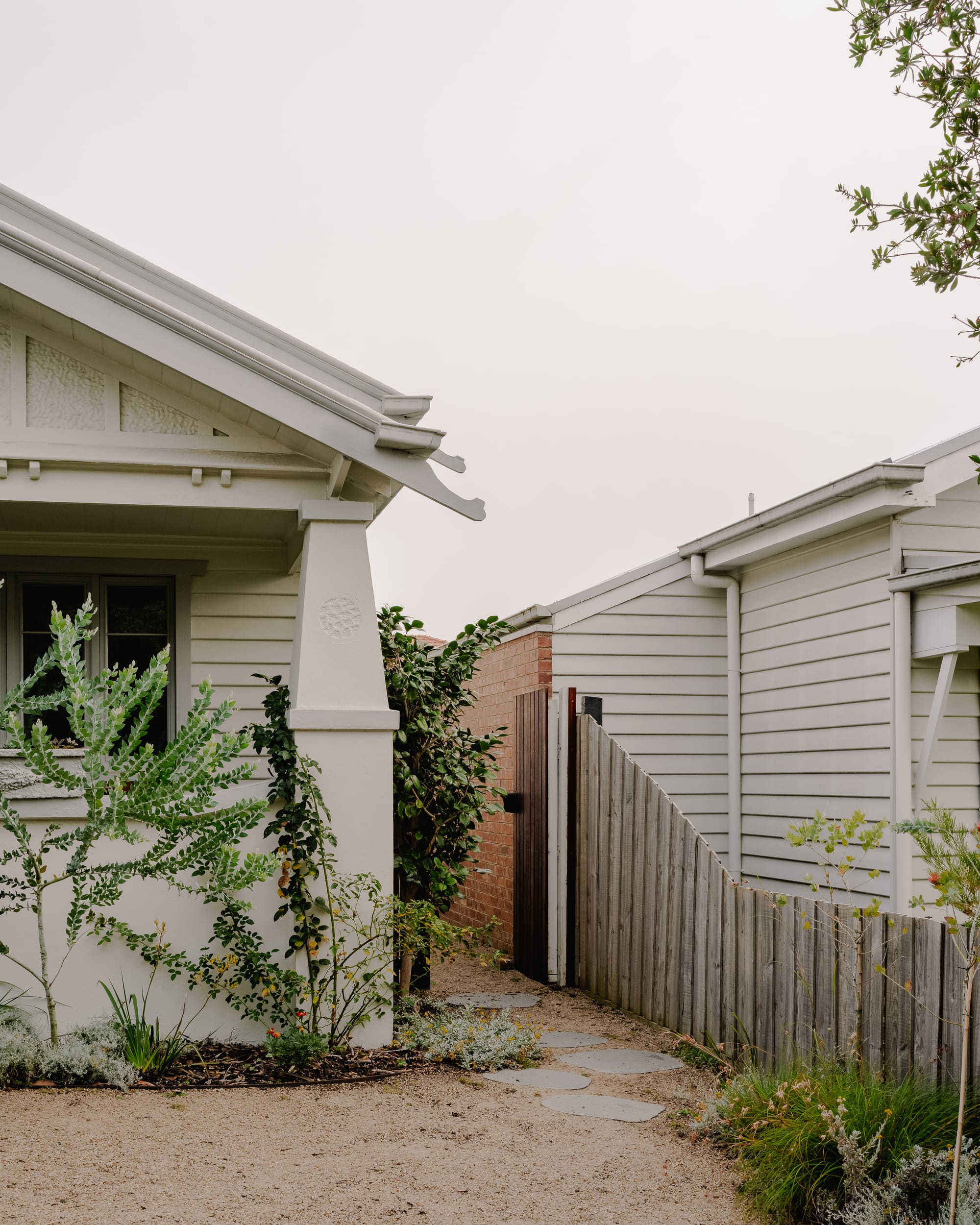House in Preston by Ashley O'Neill Architects. Photography by Victor Vieaux. Facade of bungalow-style home with enclosed porch to the left, with a paved footpath leading down the side of the house and alongside a red brick wall.