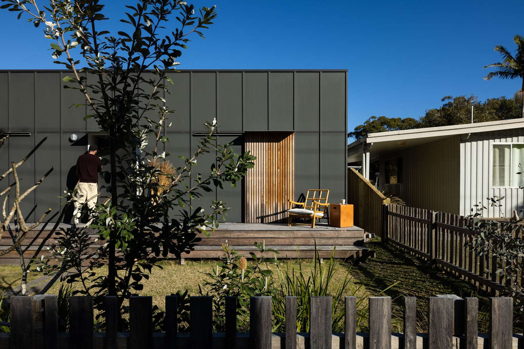 Drip Dry House by Marker Architecture. Photography by Simon Whitbread. Dark green metal clad modern granny flat in backyard, with small timber stepped deck, grass front yard and weathered picket fence.