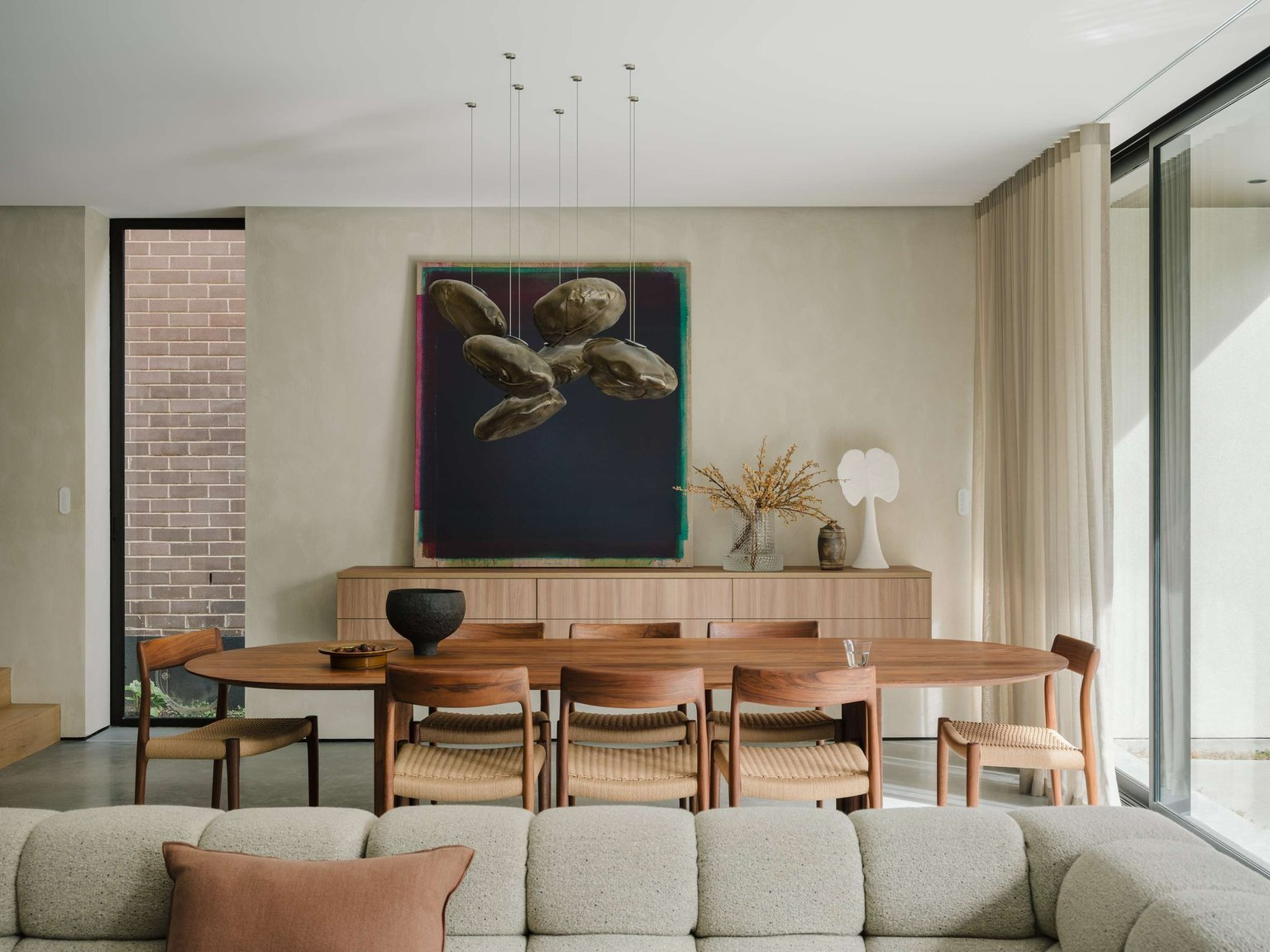 Petersham House by Cateaux Architects. Photography by Clinton Weaver. Open plan dining and living space with grey couch in foreground, long round timber dining table with rattan chairs in midground and a limewashed wall with timber sideboard, black wall art and several pendant lights suspended over the table.