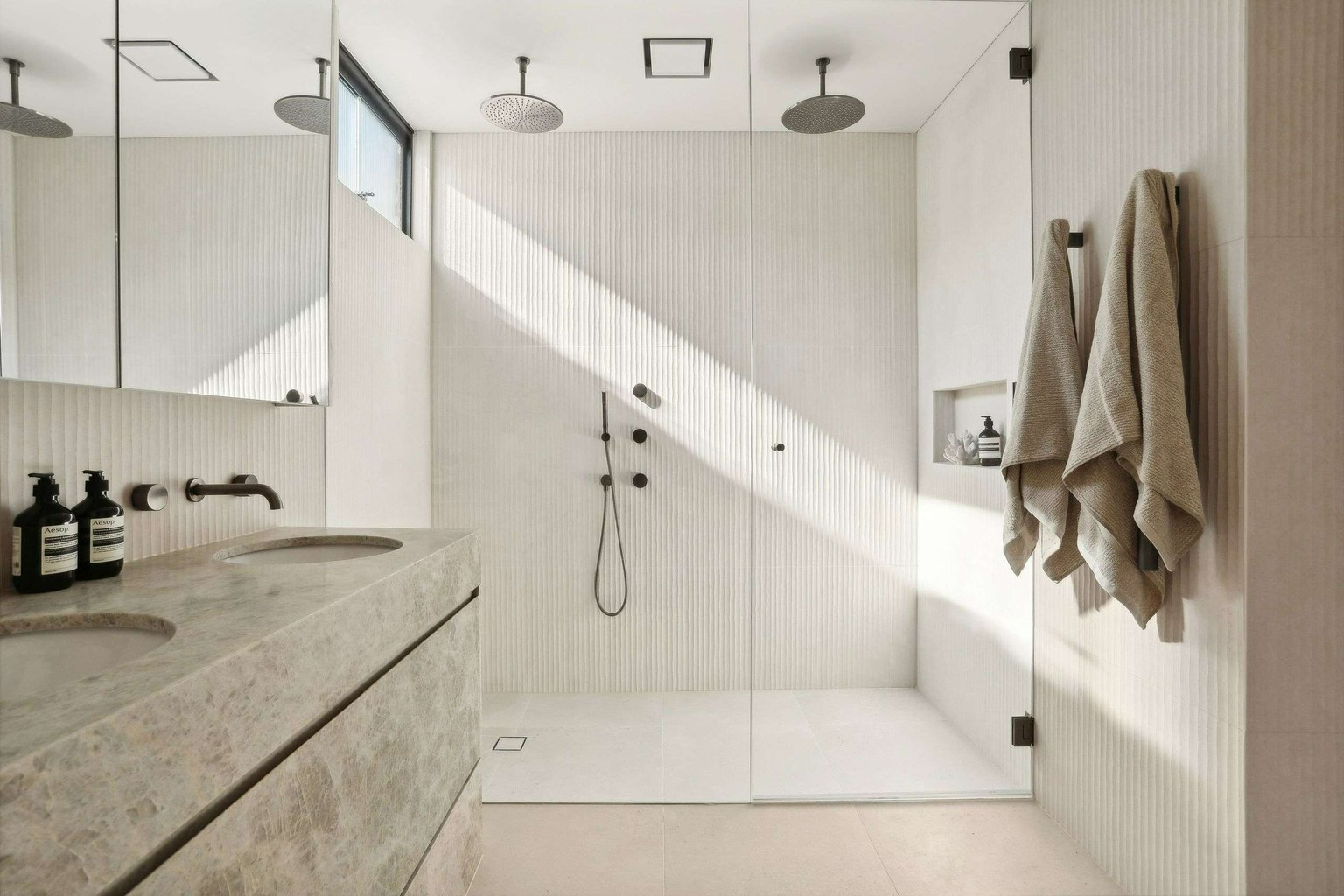 Sunset Calling, Dover Heights by BJB Architects. Photography by Core Photography Australia. Bathroom wet room with large double shower with beige wall tiles and white floor tiles and a grey marbled counter with mirror.