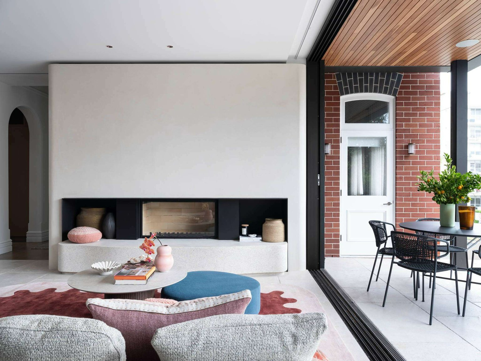 Manly 23 by Watershed Architects. Photography by Tom Ferguson. Living room with large integrated fieplace, colourful furnishings and a full-length sliding door opening onto a red brick and paved patio area with black outdoor furniture.