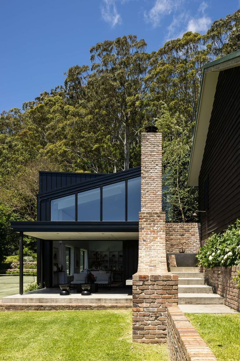 Wicklow Hills by Robertson Collectif. Photography by Simon Whitbread. Rear facade of multistorey home with brick and black clad exterior, pitched roof with window fittings and concrete stairs leading into the house from a grassed backyard.