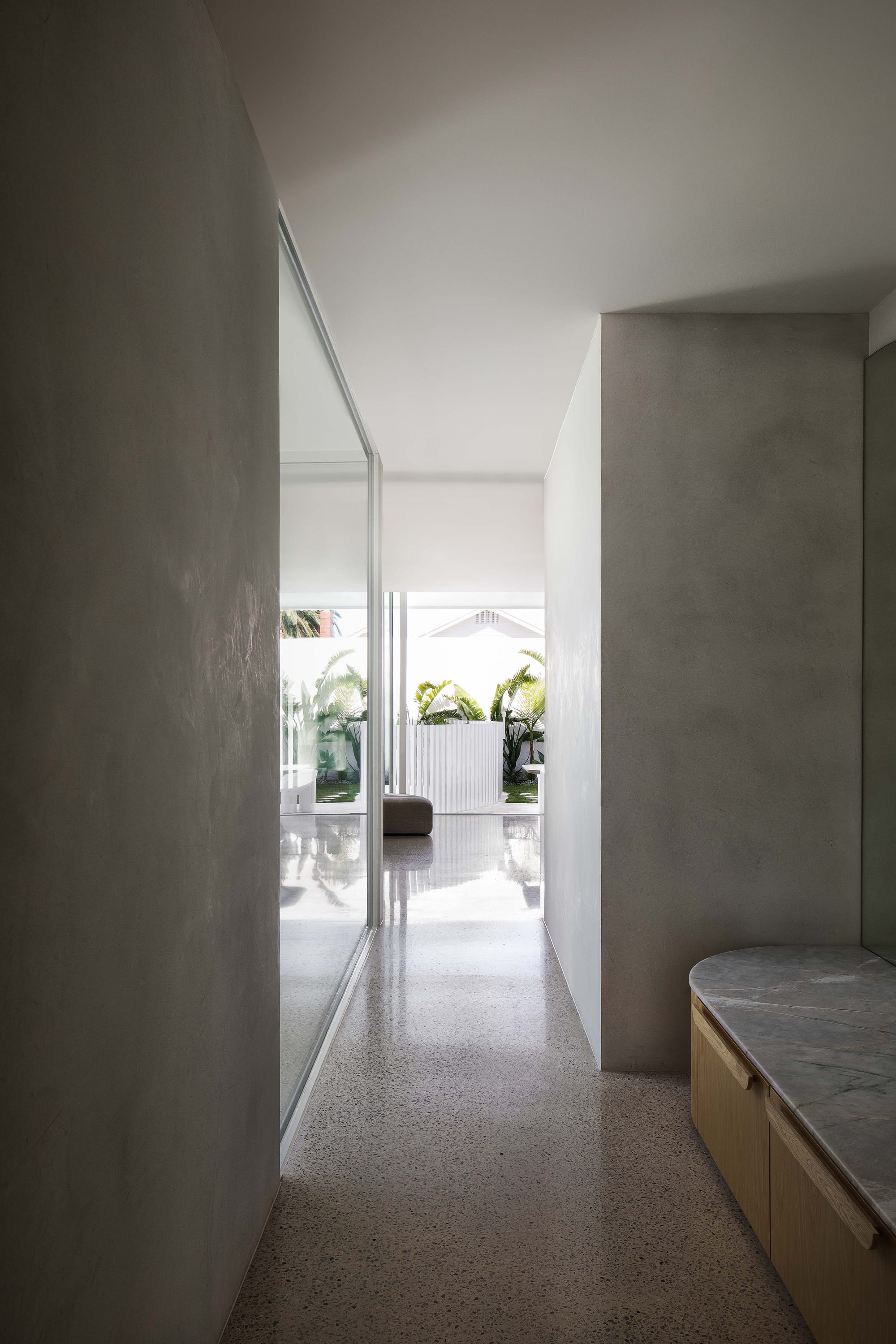 Alba House by Taouk Architects. Photography by Emily Bartlett. Hallway with polished concrete floors, limewashed walls, integrated bench seating in timber and stone and full height glass doors opening onto outdoor areas.
