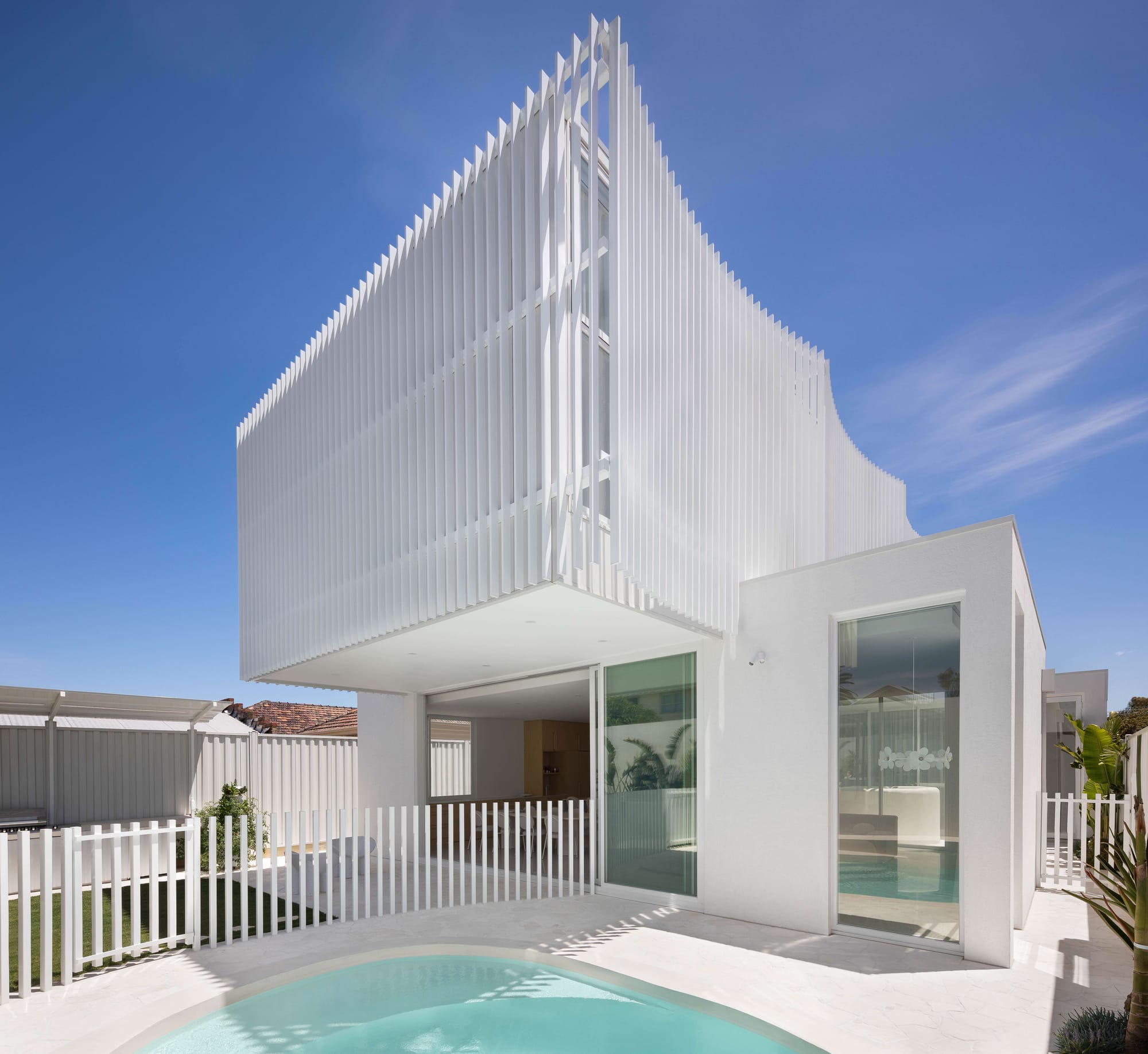 Alba House by Taouk Architects. Photography by Emily Bartlett. Rear facade of two story residential home with in-ground pool white tiled flooring, large second story overhand with fluted metal exterior and large glass windows connecting ground floor and pool area.