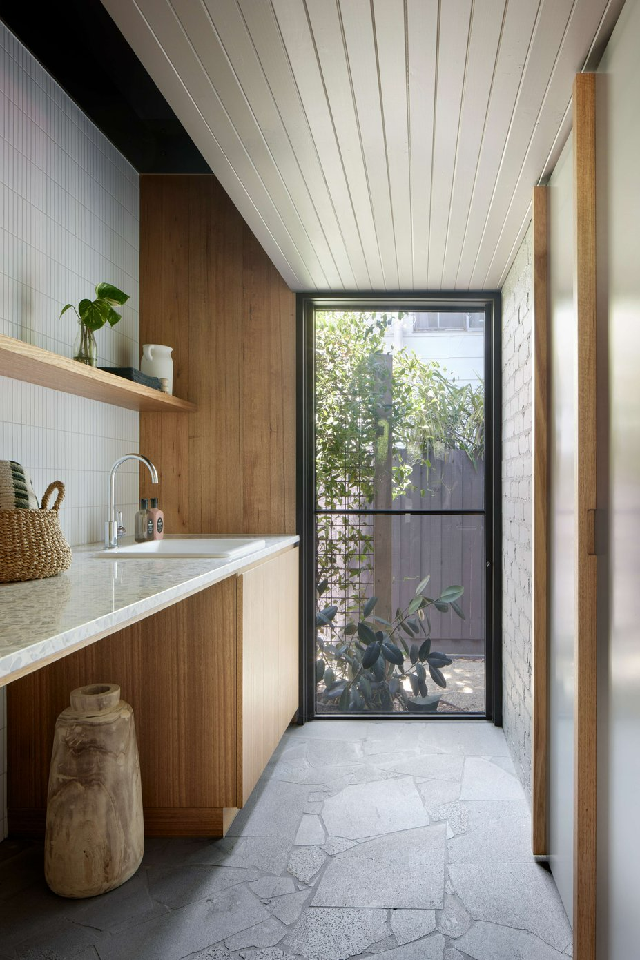 Eaglemont House by BENT Architecture. Photography by Tatjana Plitt. Laundry with timber countertops, stone paver flooring, full height window looking onto garden and stone countertop with white mosaic splashback.