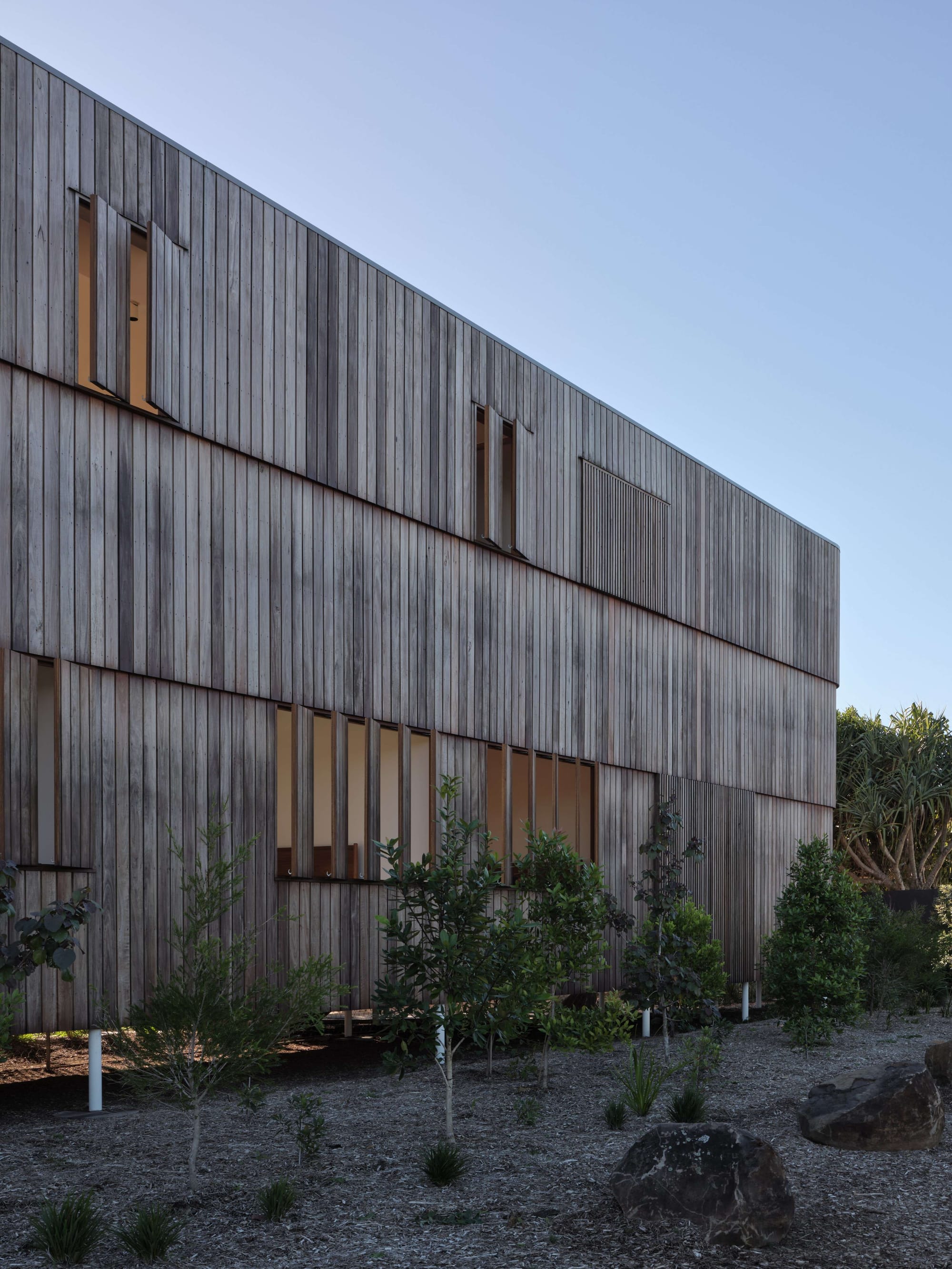Blok Peregian by Blok Belongil. Photography by Christopher Frederick Jones. Facade od double storey home with weathered timber clad exterior, timber louvre windows and native garden with trees and mulch.
