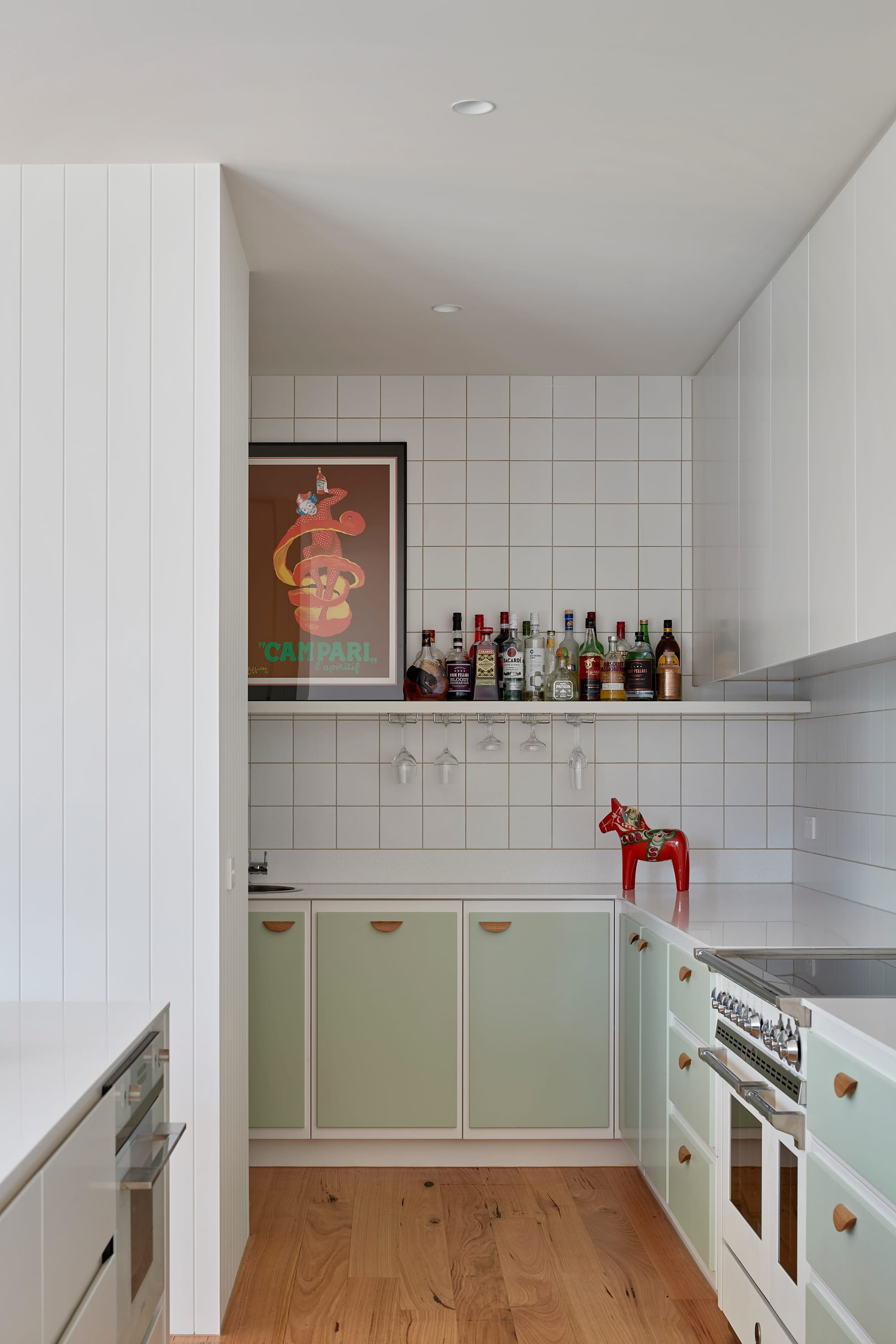 The Blairgowrie Beach Club by Bower Architecture & Interiors. Photography by Shannon McGrath. A small bar with liquors in a pantry space with white square tiles and mint green colored furnitures.