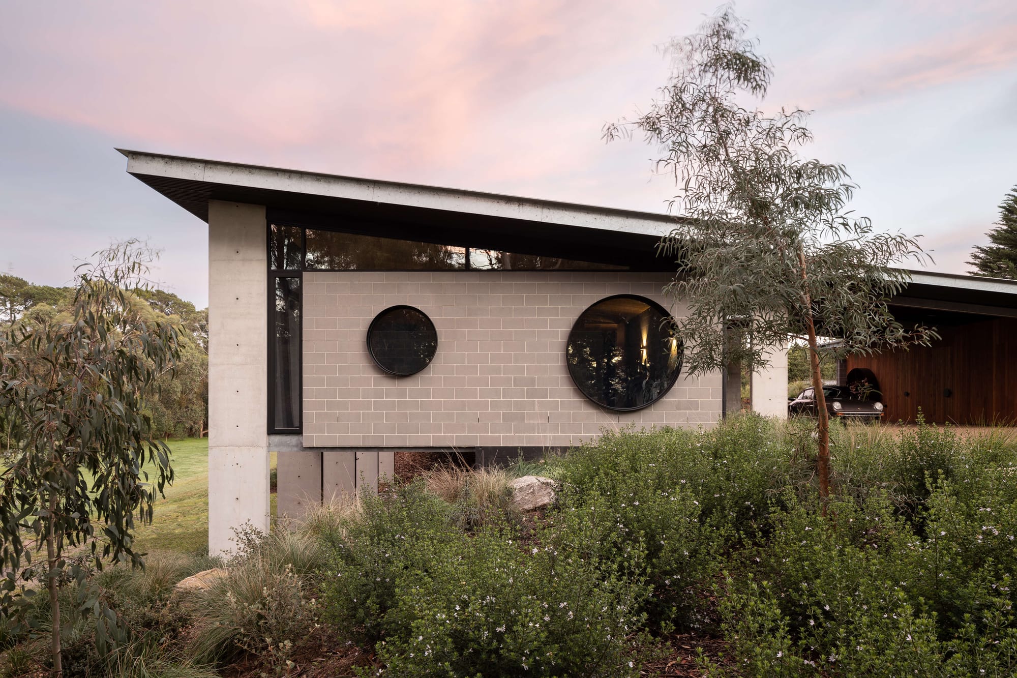 Merricks House by Aktis Architects. Photography by Timothy Kaye. Modern house with angled roof, concrete and blockwork walls, and two circular windows set against a lush native garden at dusk.