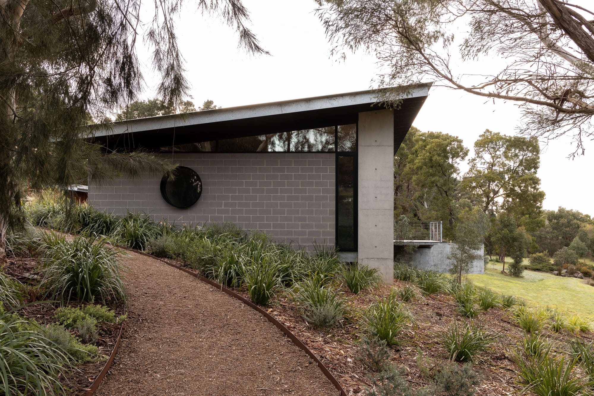 Merricks House by Aktis Architects. Photography by Timothy Kaye. Side view of a modern home with circular window, grey blockwork, and angled roof, bordered by a winding gravel path and native landscaping.