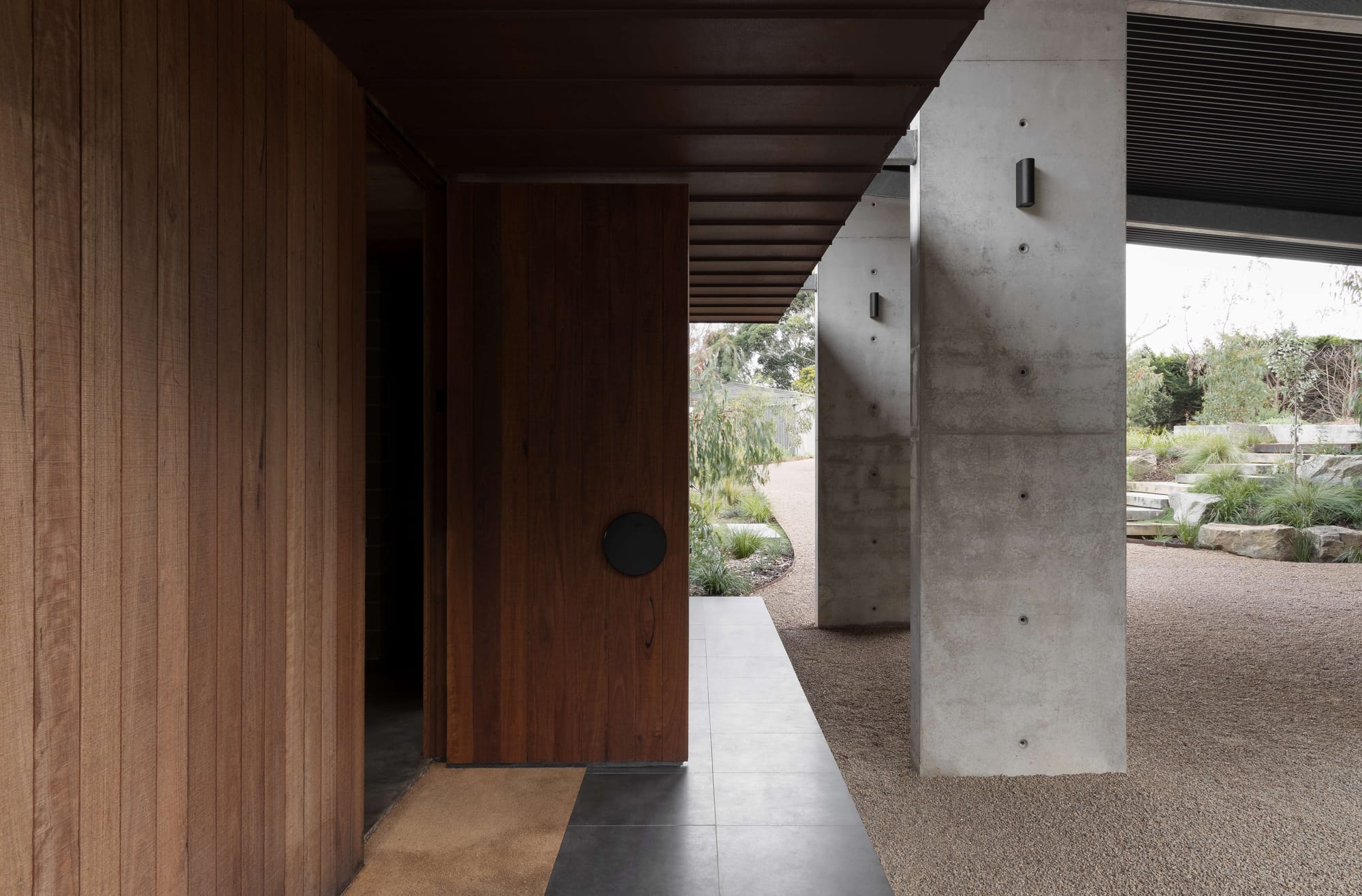 Merricks House by Aktis Architects. Photography by Timothy Kaye. Entryway detail featuring timber cladding, recessed doorway with circular handle, and exposed concrete columns framing a gravel path.