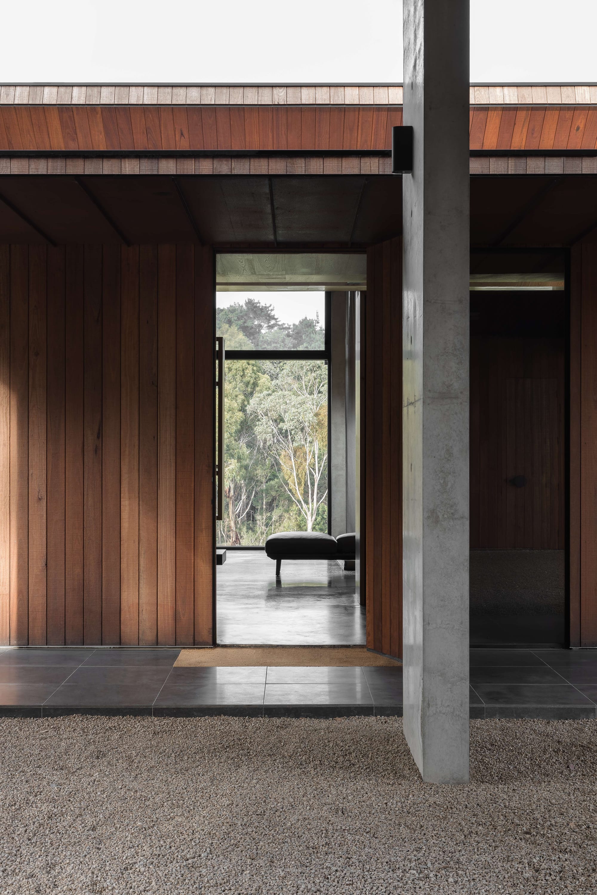 Merricks House by Aktis Architects. Photography by Timothy Kaye.Framed view through timber-clad entry and concrete column to a bench seat and forest beyond, with gravel and tiled flooring.