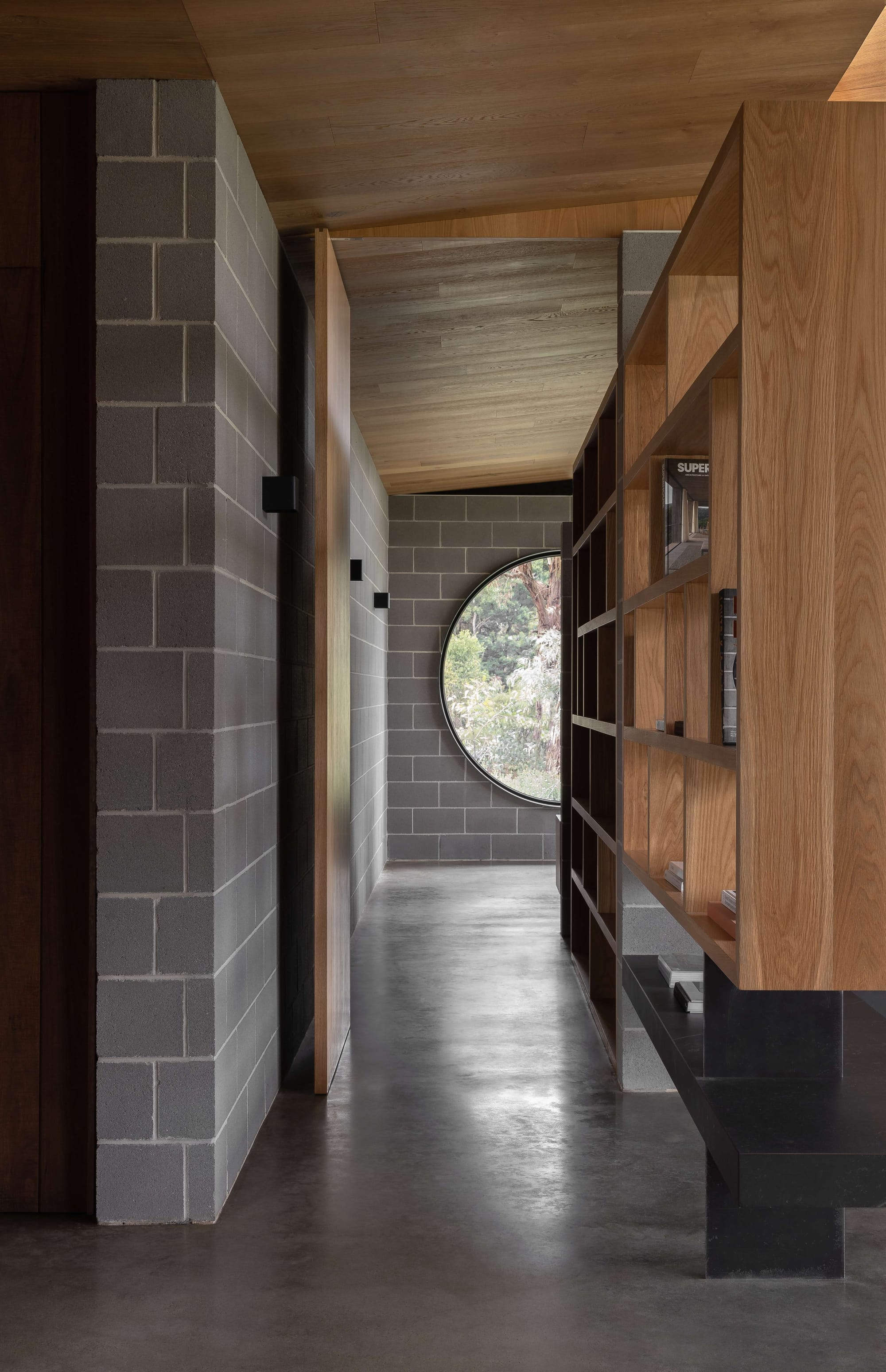 Merricks House by Aktis Architects. Photography by Timothy Kaye. Interior hallway with concrete block walls, timber ceiling, floating bookshelves, and a large circular window framing a garden view.