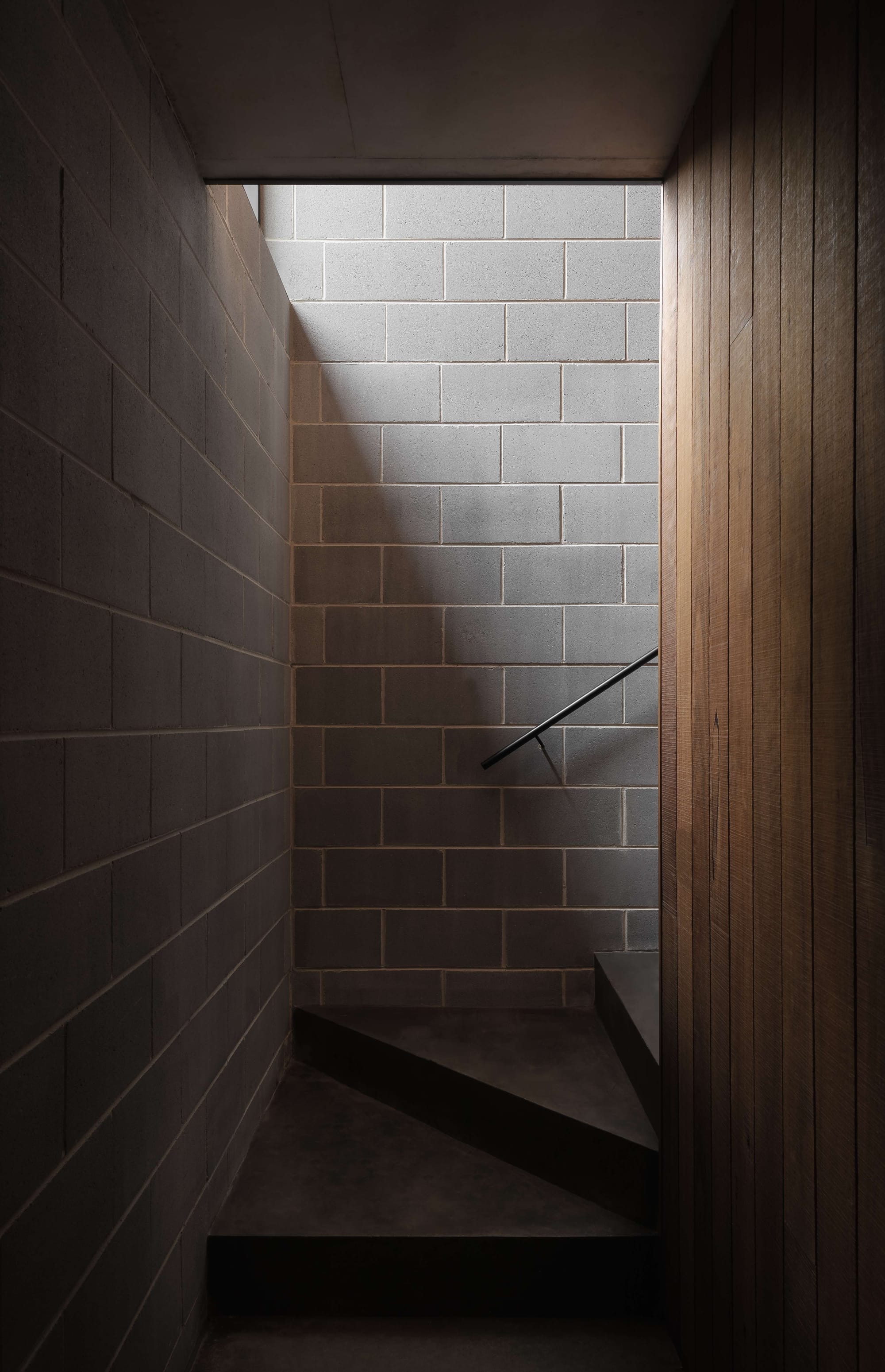 Merricks House by Aktis Architects. Photography by Timothy Kaye. Minimal stairwell with grey concrete block walls, timber cladding, and soft natural light casting shadows across the textured surfaces.