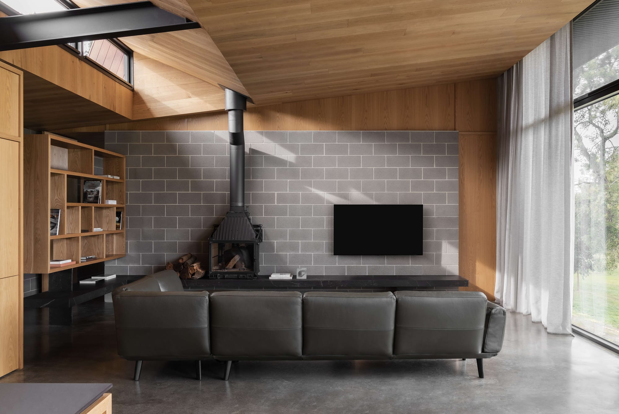 Merricks House by Aktis Architects. Photography by Timothy Kaye. Modern living room with concrete block feature wall, black wood-burning stove, leather sofa, timber shelving, and floor-to-ceiling windows.
