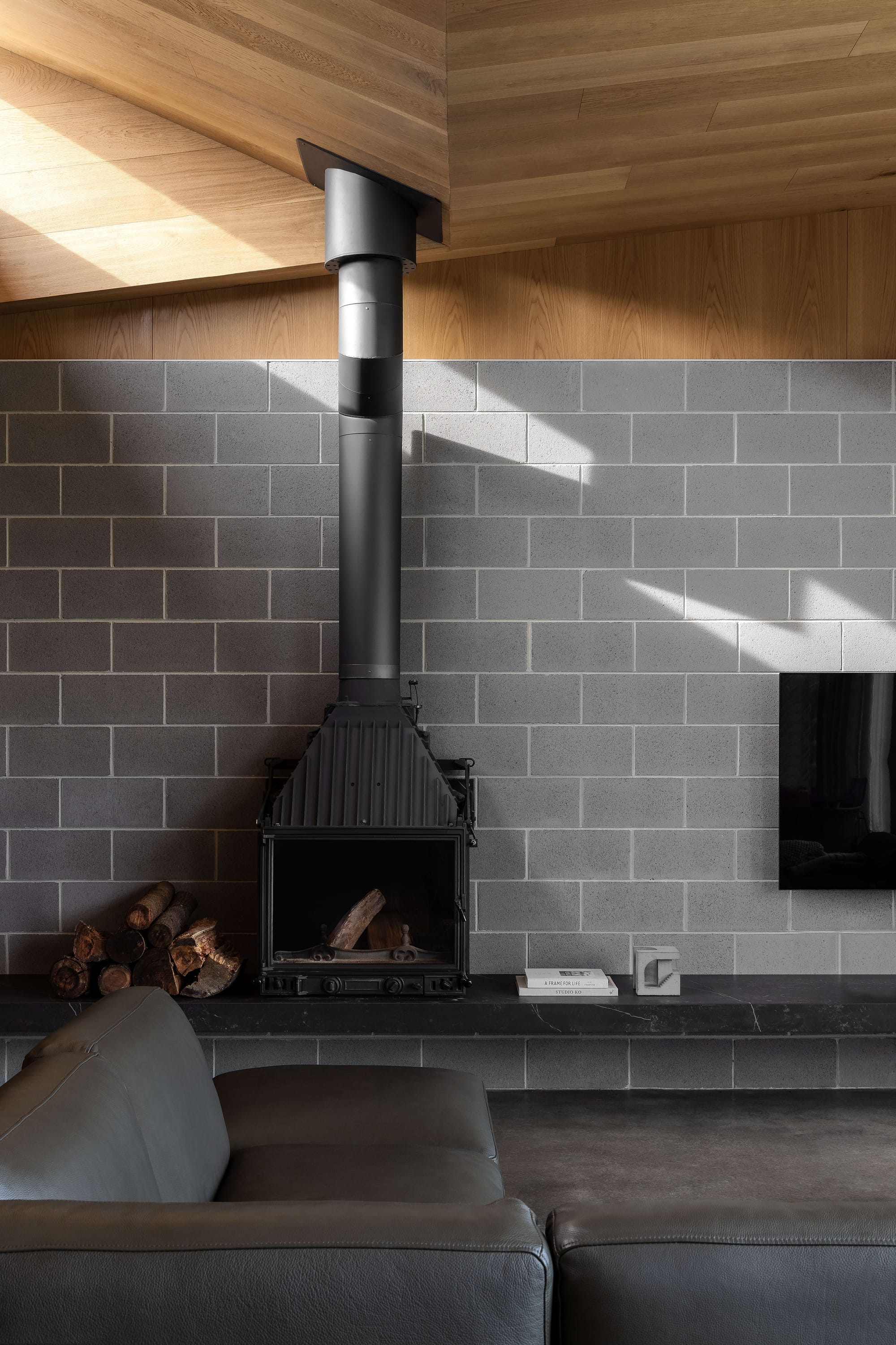 Merricks House by Aktis Architects. Photography by Timothy Kaye. Close-up of a modern wood-burning fireplace with a black flue, set against a grey blockwork wall and timber ceiling, with stacked firewood and minimalist decor.