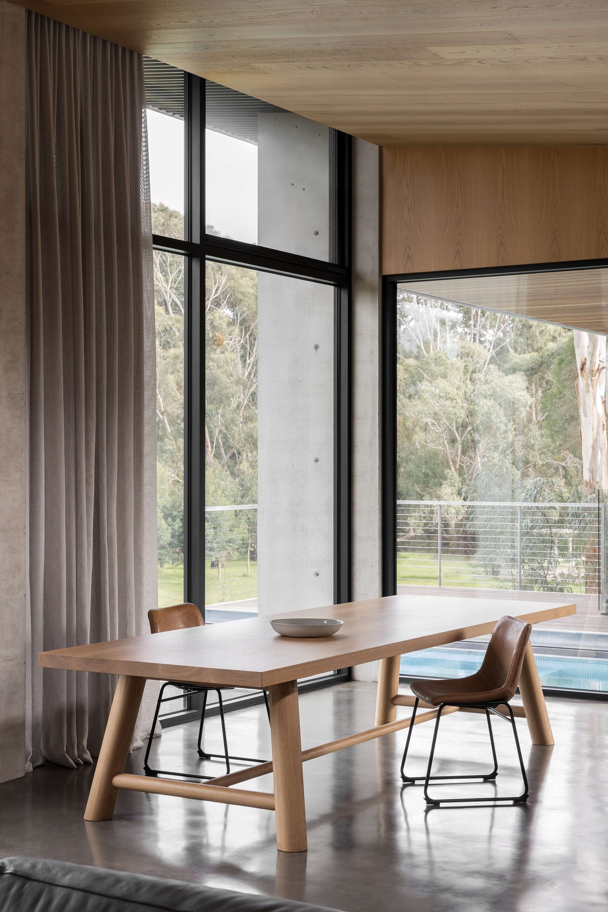 Merricks House by Aktis Architects. Photography by Timothy Kaye. Dining area with a solid timber table, leather chairs, full-height windows, and views to a landscaped garden and pool beyond.