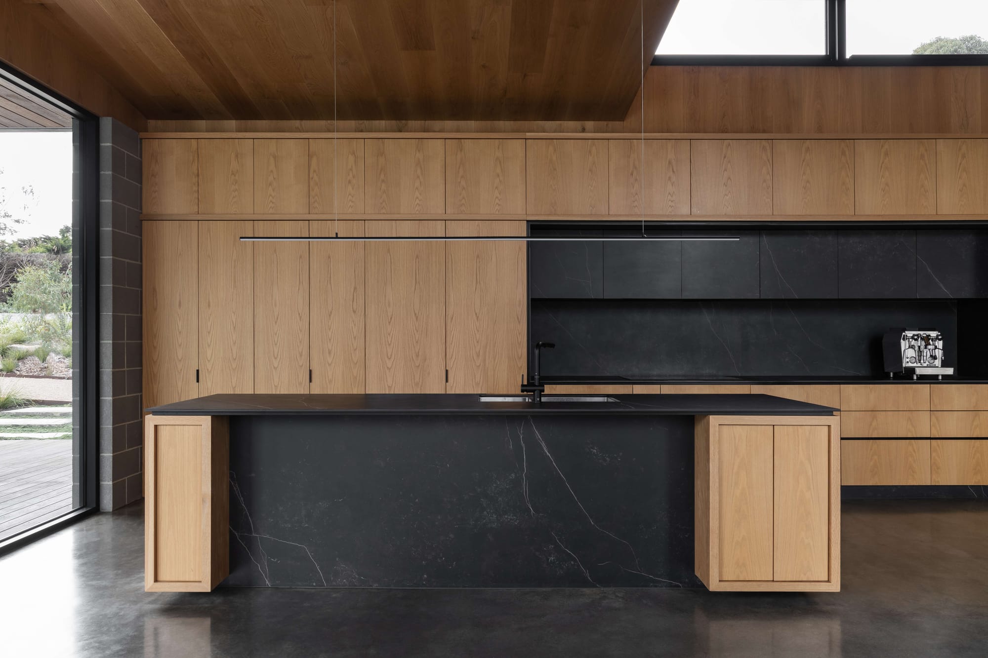 Merricks House by Aktis Architects. Photography by Timothy Kaye. Contemporary kitchen with black marble island, timber cabinetry, minimalist pendant lighting, and polished concrete floors.